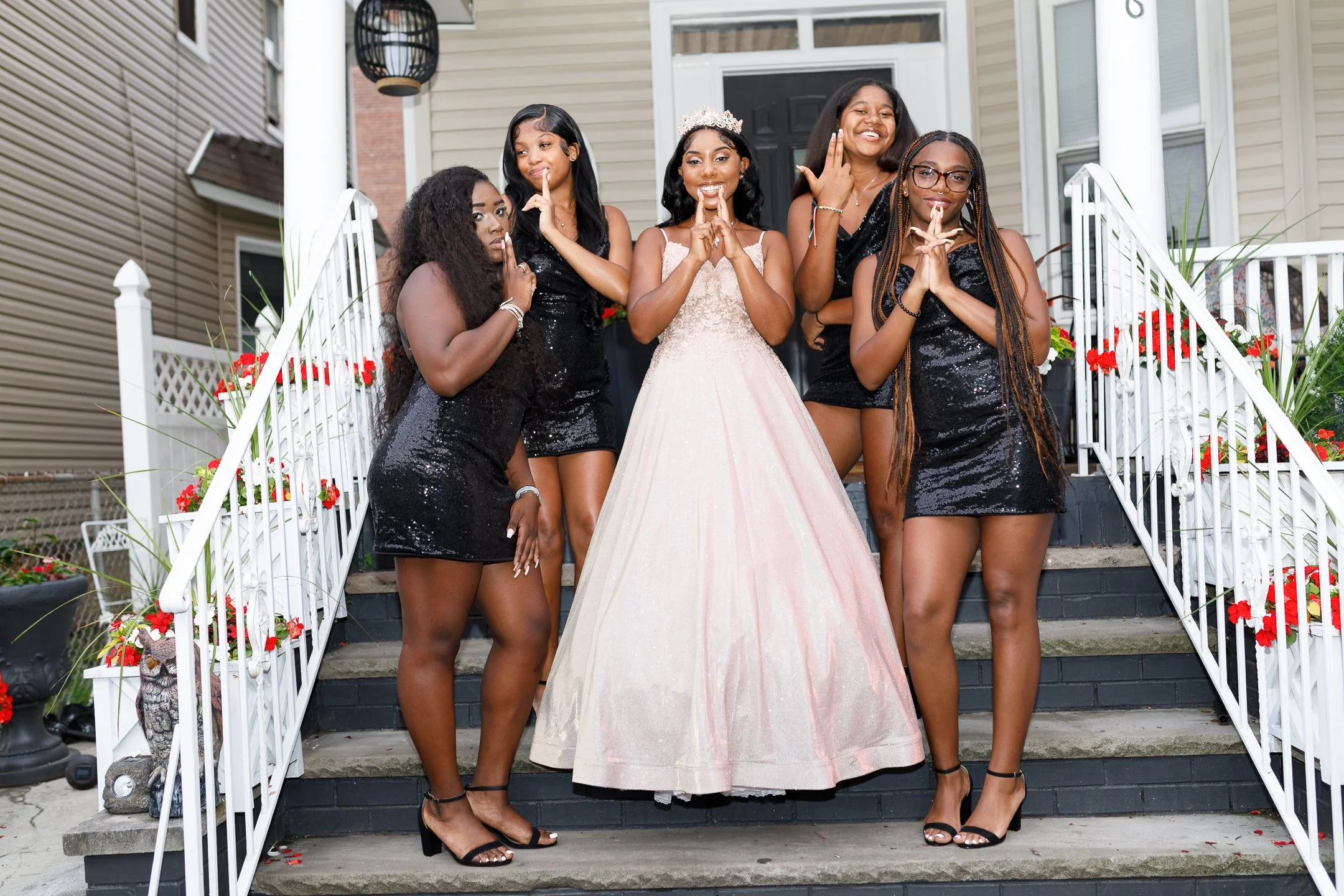 A bride in a pink ball gown stands on the front steps of a house, surrounded by five women in black dresses. The women are posing with finger over lips, some smiling and some serious. The scene is decorated with red flowers on the railings.