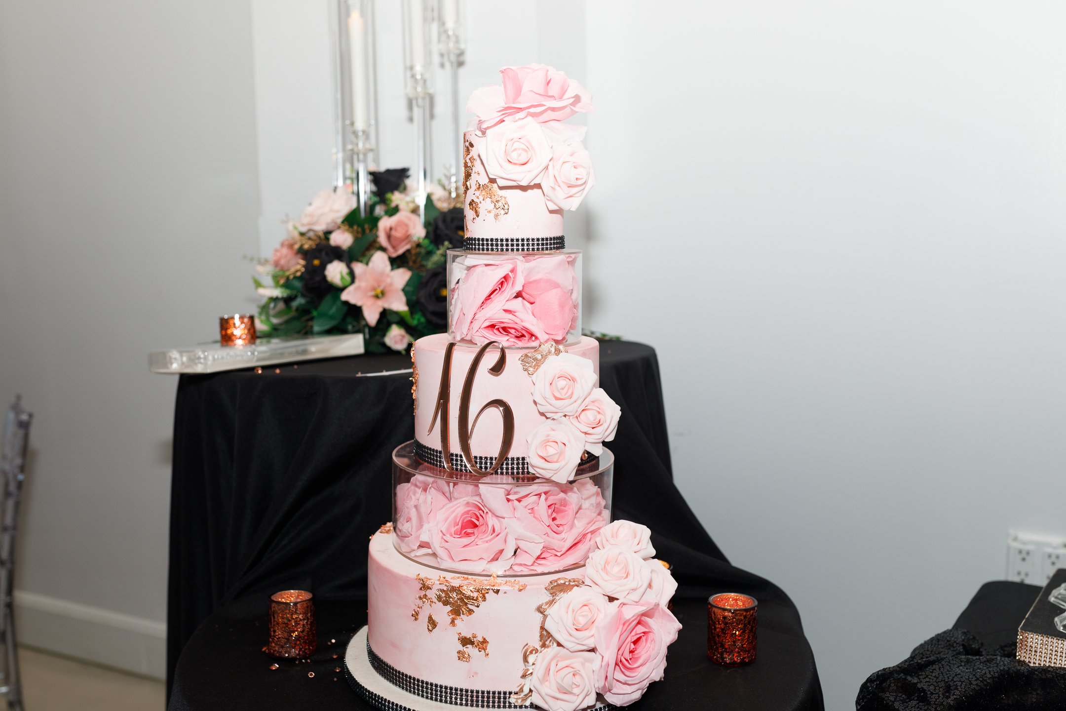 Pink and white five-tiered wedding cake decorated with pink roses, gold accents, and black ribbon, with the number 16 on the second tier, set on a black table with floral arrangements and decorative candles in the background.