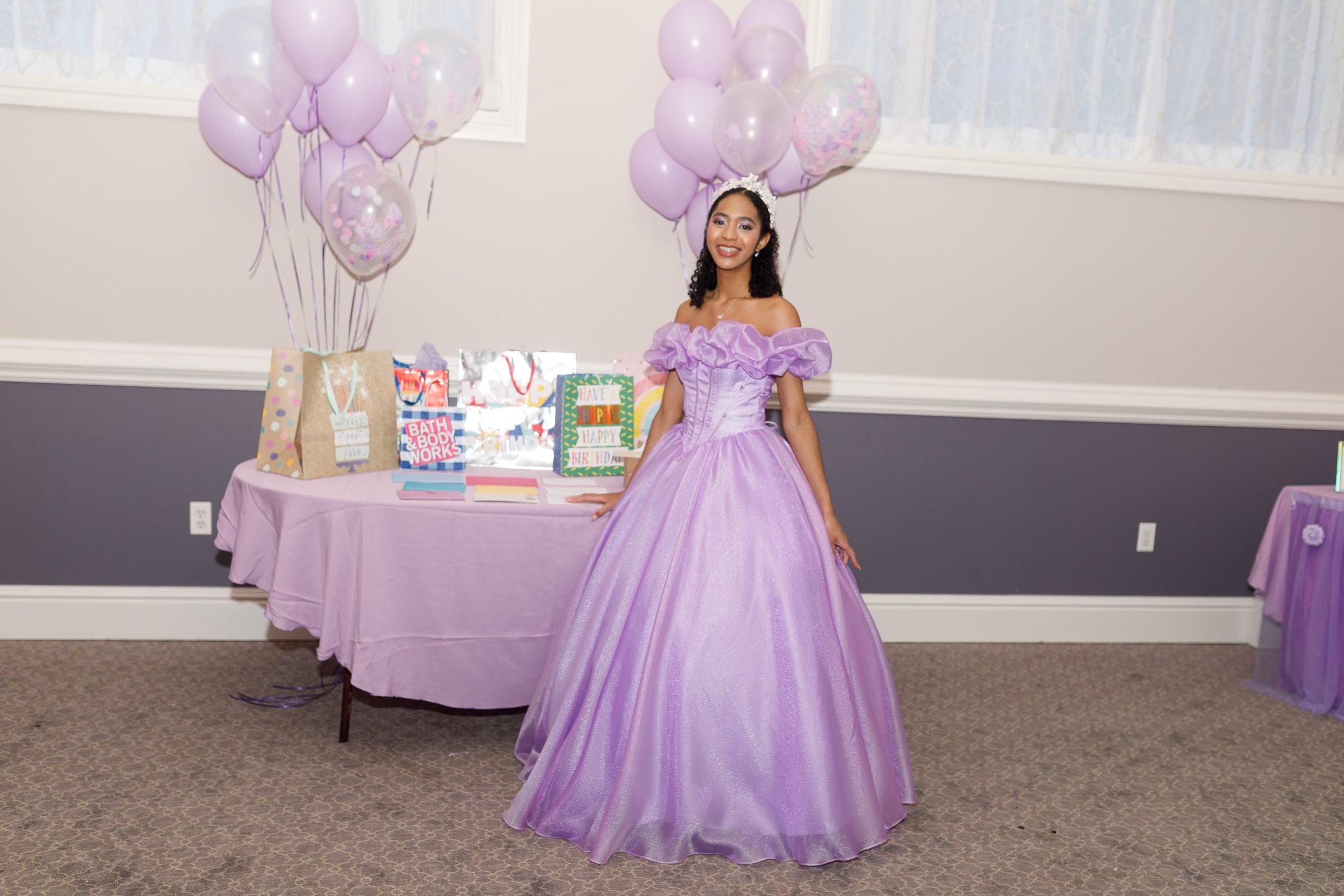 Young woman in a lavender ball gown with puffed sleeves and a tiara, smiling at a celebration with balloons and gift bags on a table.