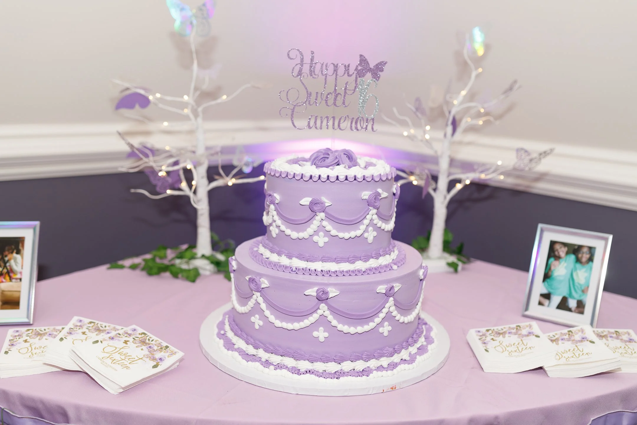 A three-tiered purple birthday cake decorated with white icing and swirls, set on a pink tablecloth with photo frames, pamphlets, and white decorative trees in the background, with a glittery topper reading 'Happy Sweet 6 Cameron'.