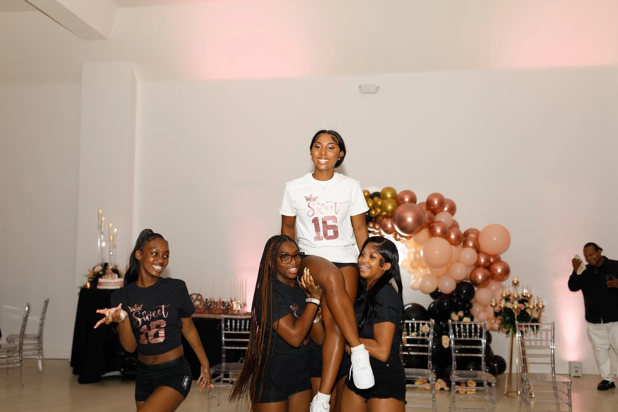 Four women celebrating at a party with balloons and decorations. One woman is lifted by three others, smiling and wearing a white shirt with the number 16. Two women are wearing black tops with similar designs and one has long braids. A man is in the