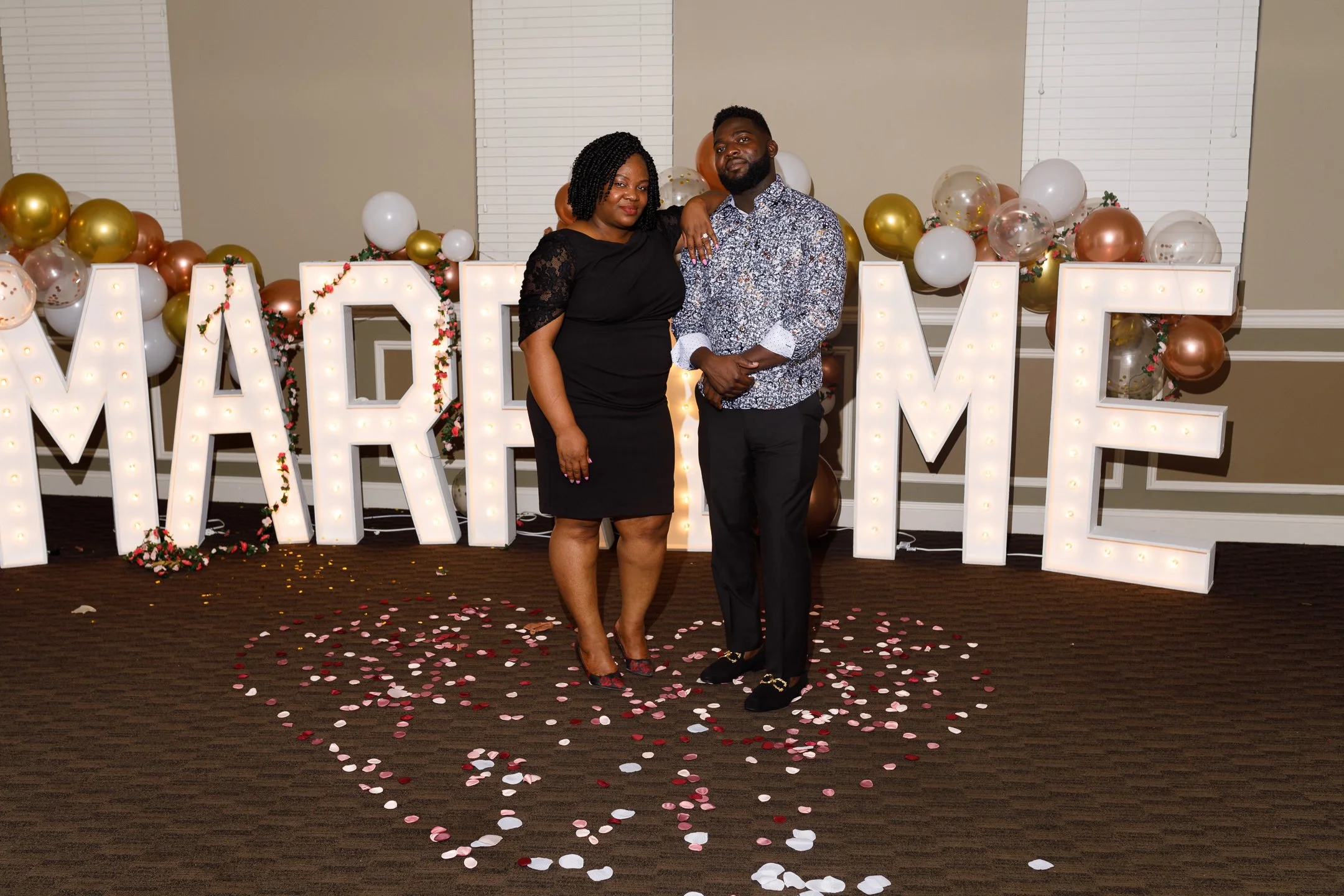 A couple standing together at a celebration with large light-up letters spelling 'MARRY ME' in the background decorated with balloons and confetti on the floor.