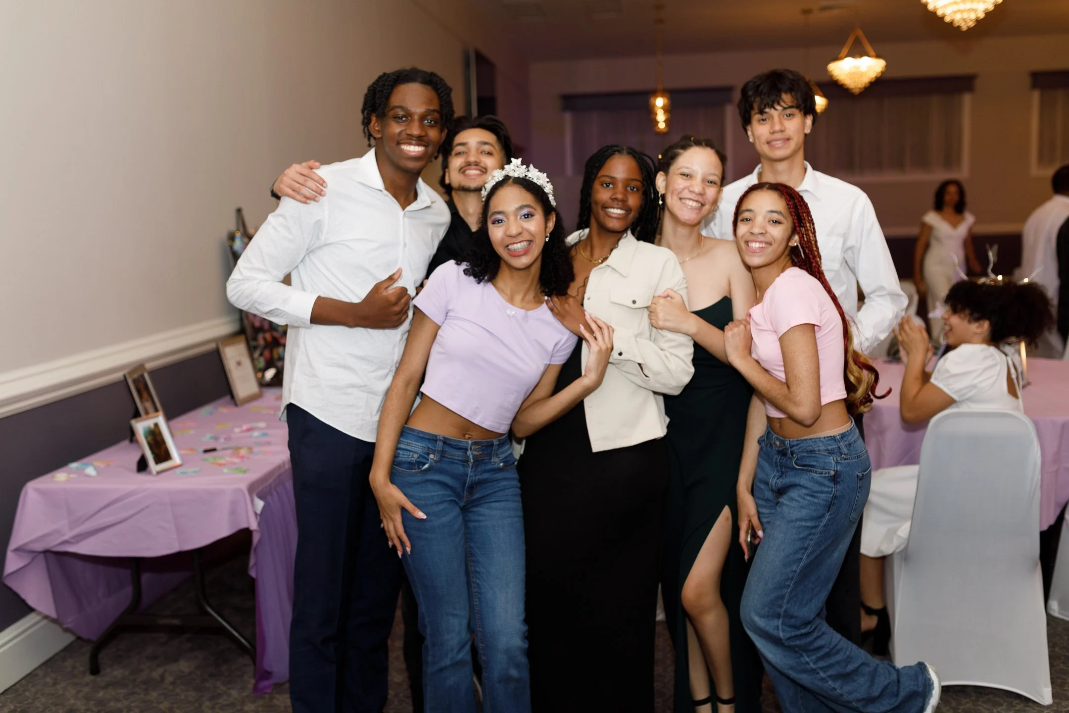 Group of eight diverse friends smiling and posing together at a party, with decorated tables and other guests in the background.