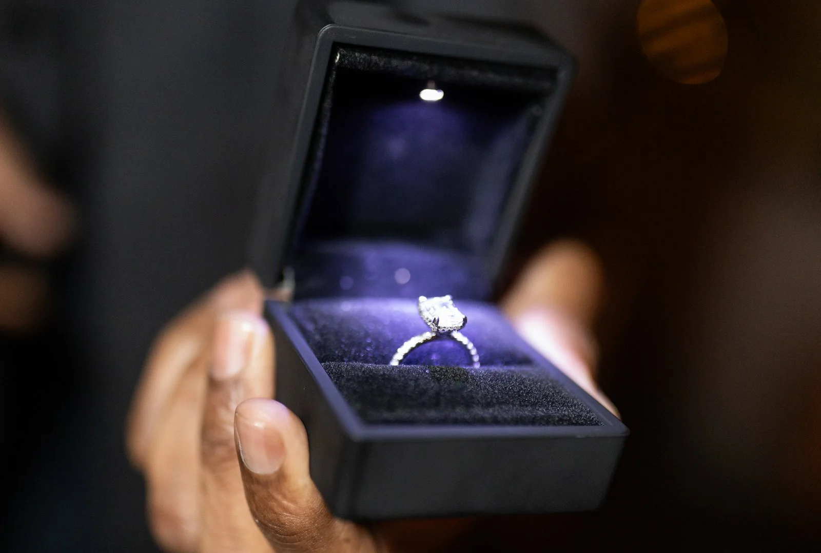 Person holding an open jewelry box with an engagement ring inside, illuminated by a small light.
