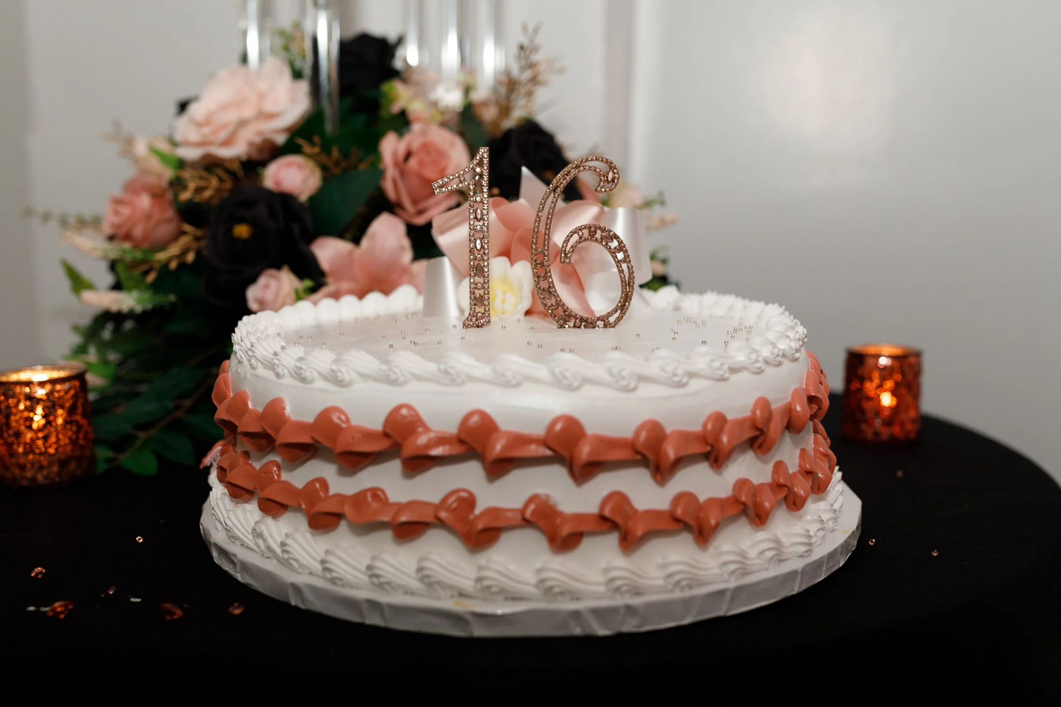 A white, two-tiered birthday cake with pink and white frosting decorations, topped with glittery gold '16' candles, and surrounded by pink and black flowers and orange candles on a black table.