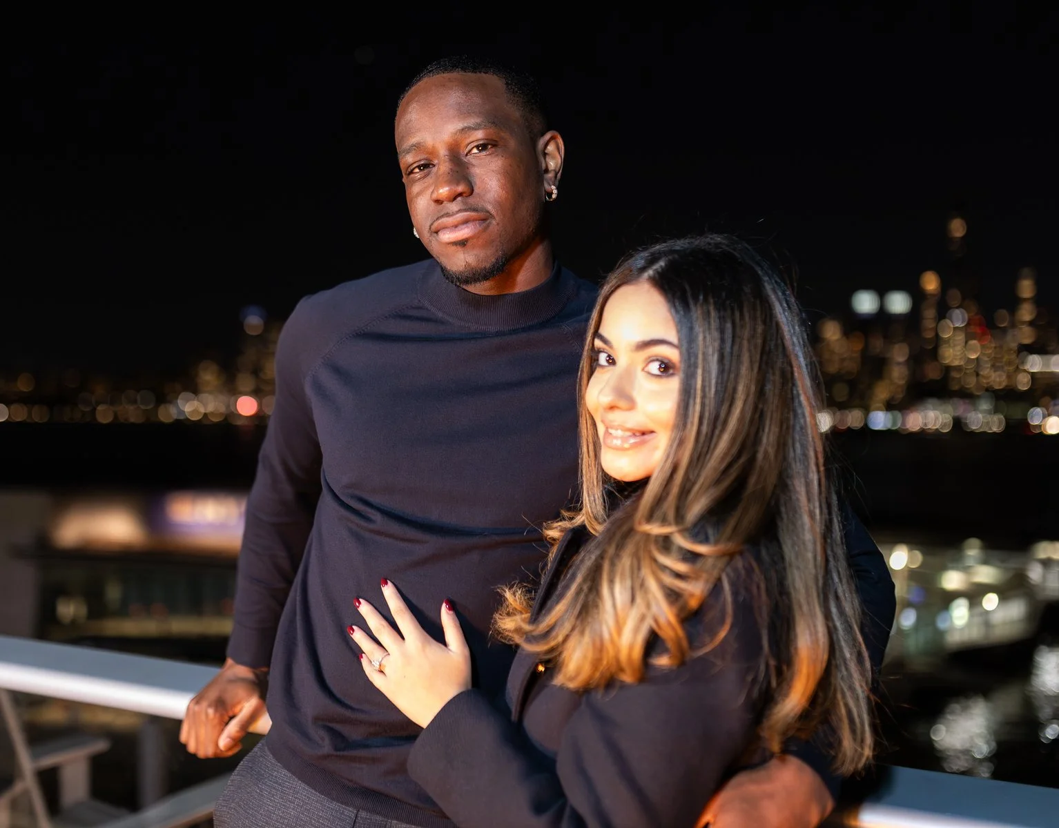A man and woman pose together on a rooftop at night, with city skyline lights in the background. The man is wearing a dark sweater, and the woman has long, dark hair with light brown tips and is wearing a dark top. They are close together, smiling so
