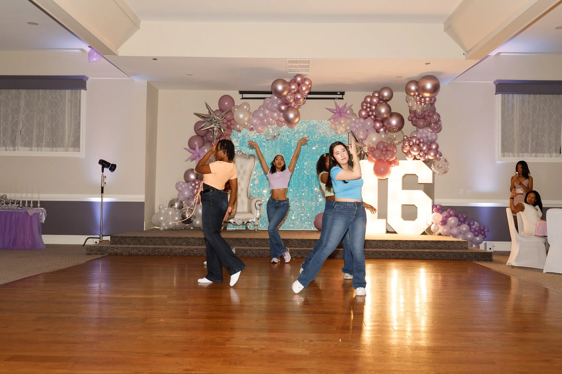 Four young women dancing on a wooden floor during a celebration with pink, purple, and silver balloons, a decorative backdrop, and a large illuminated number six.