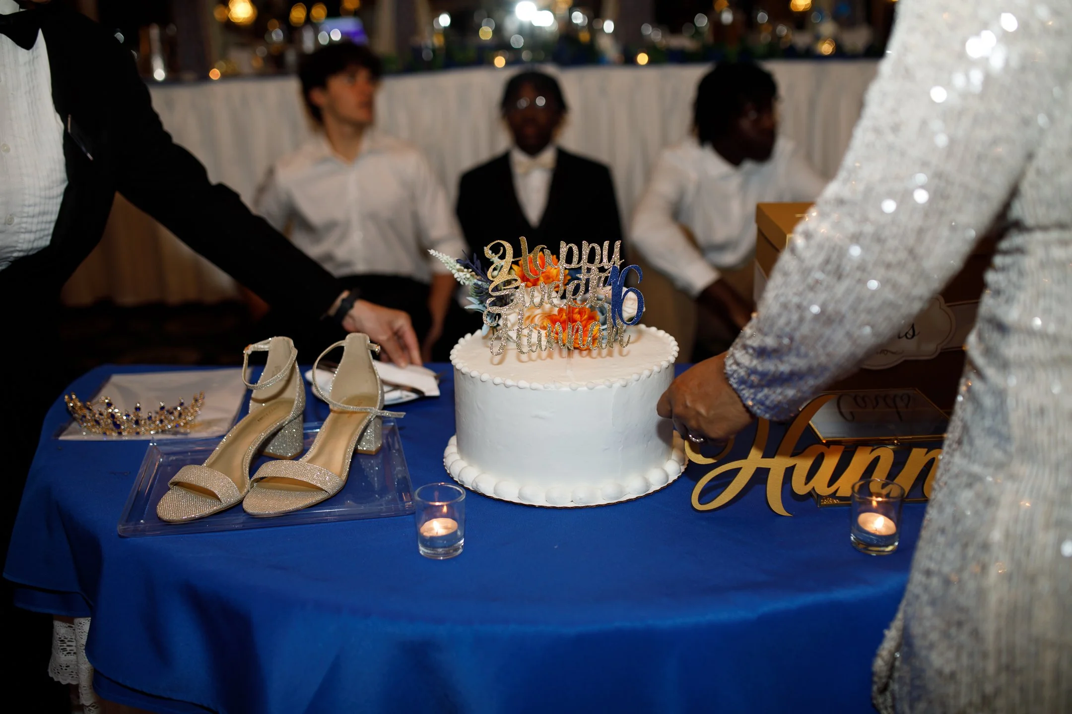 A white birthday cake with seasonal decorations on top, including a topper that says 'Happy 16th Birthday,' is on a table covered with a blue tablecloth. There are glittery gold high-heeled shoes and a tiara on display next to the cake. Two women are