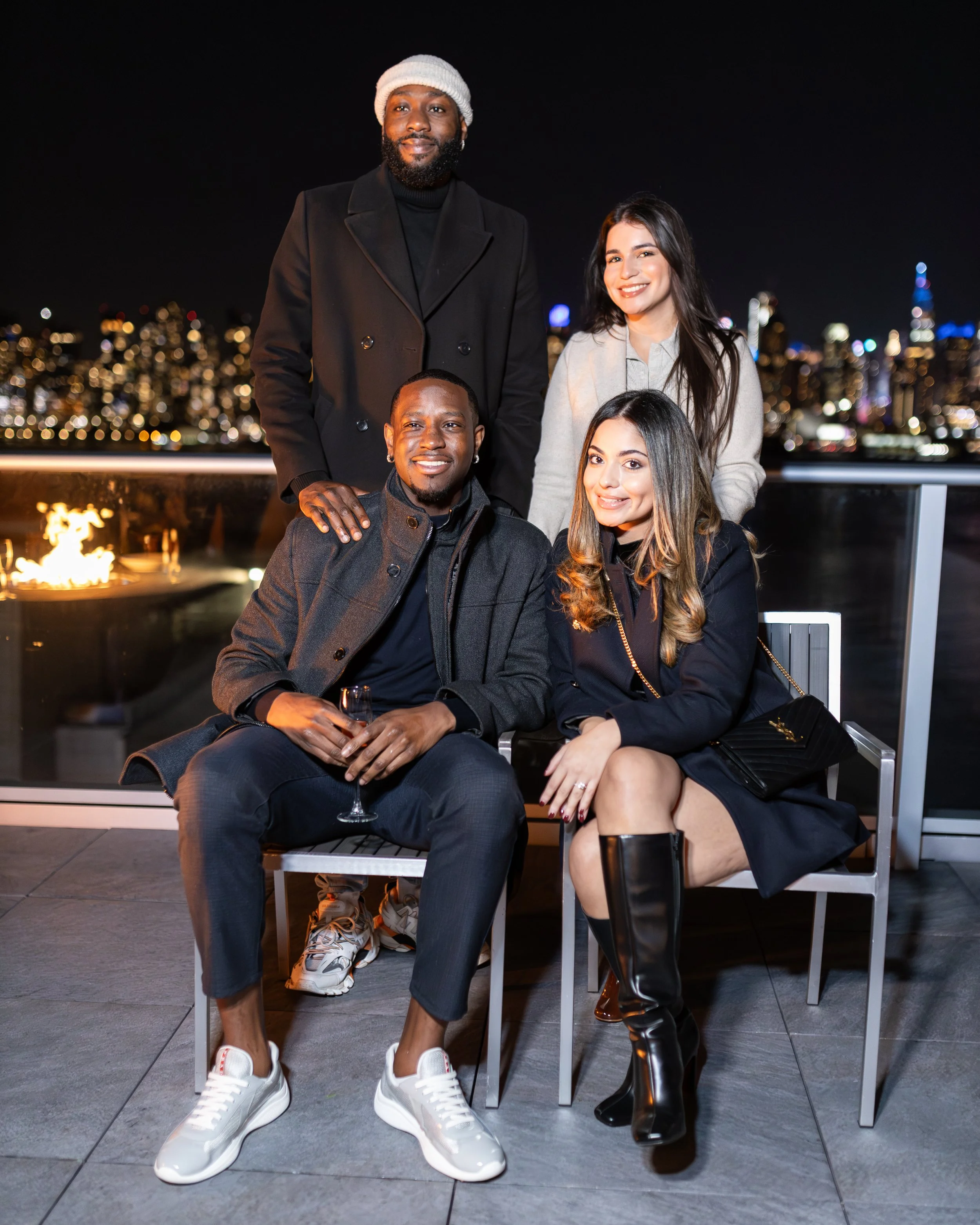 Group of five friends on a rooftop at night with a city skyline in the background, two seated and three standing, smiling and dressed stylishly.