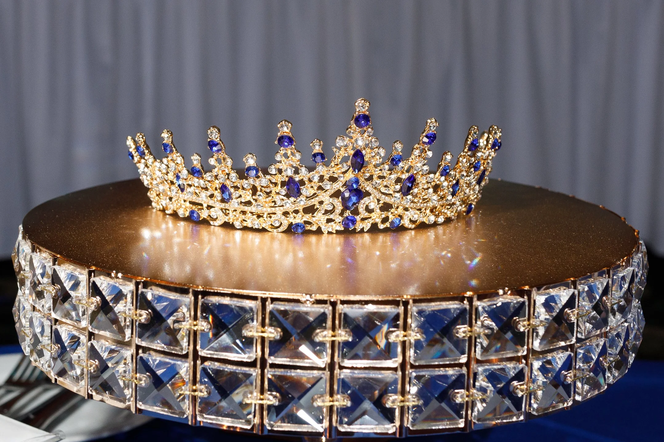 A gold tiara with blue and clear gemstones on a mirrored surface with decorative edge, against a gray curtain background.