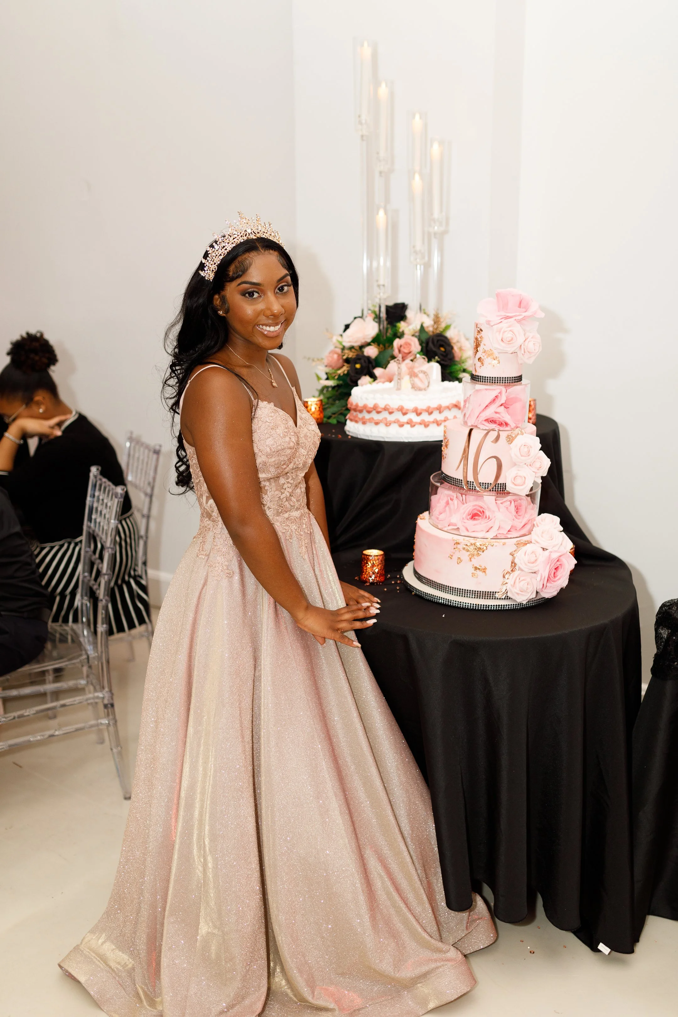 Young woman in a pink gown with a tiara standing next to a multi-tiered pink and white birthday cake with roses, celebrating her 16th birthday.