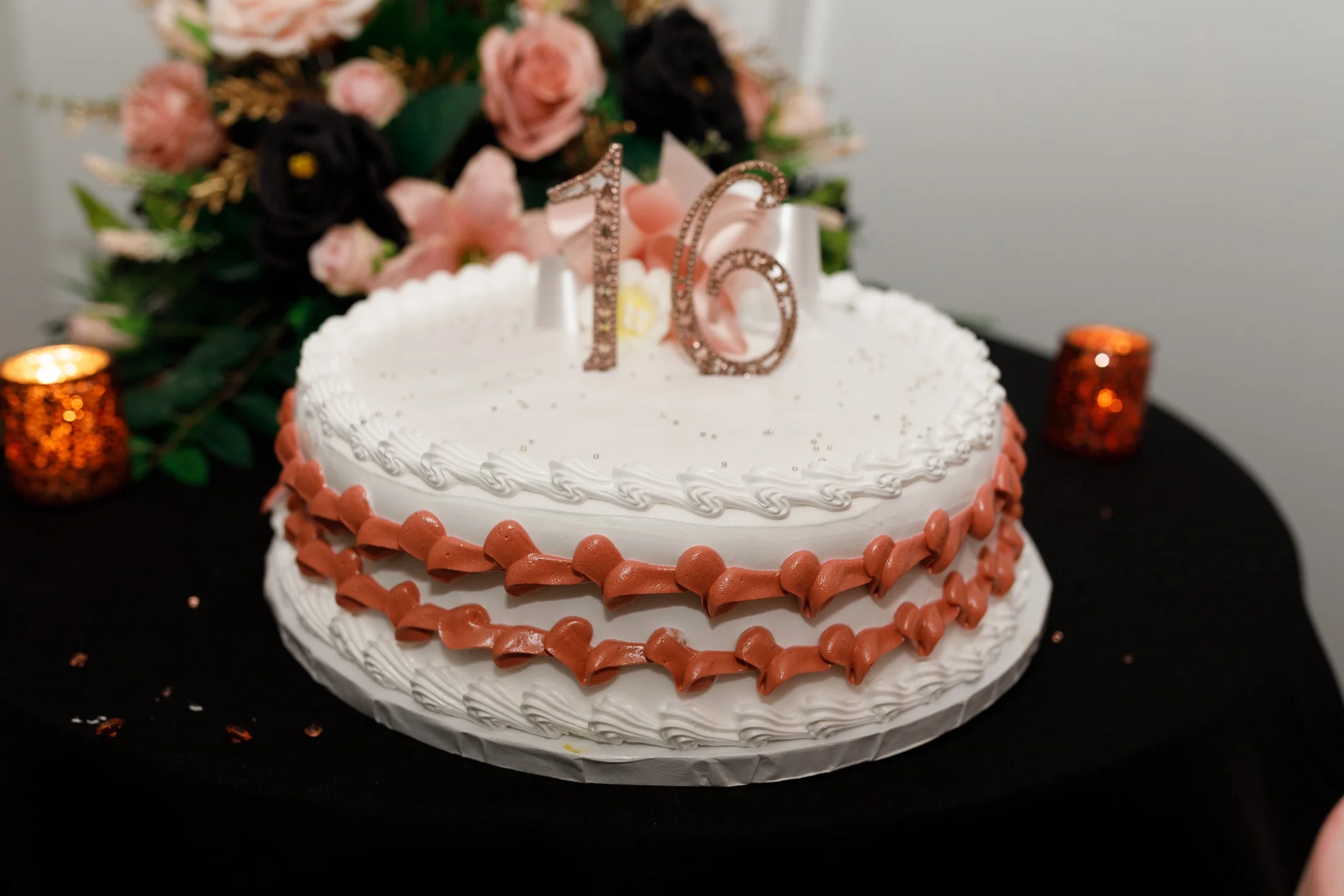 A white birthday cake decorated with pink and white icing, topped with a gold number '16' candle, with a bouquet of pink and dark purple flowers in the background, and small orange candles on a black table.