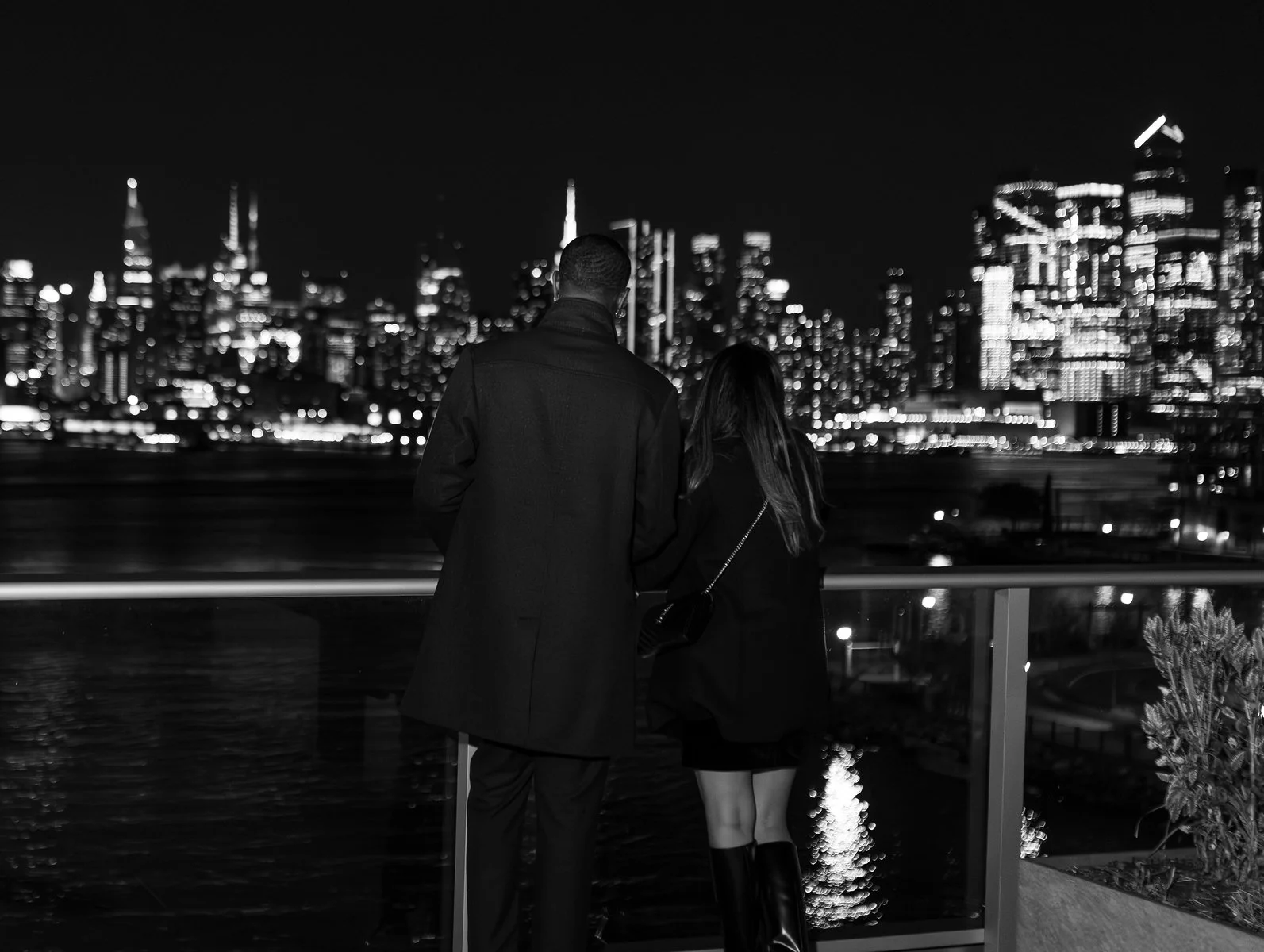 A man and woman standing together on a balcony at night, overlooking a city skyline illuminated with bright lights.