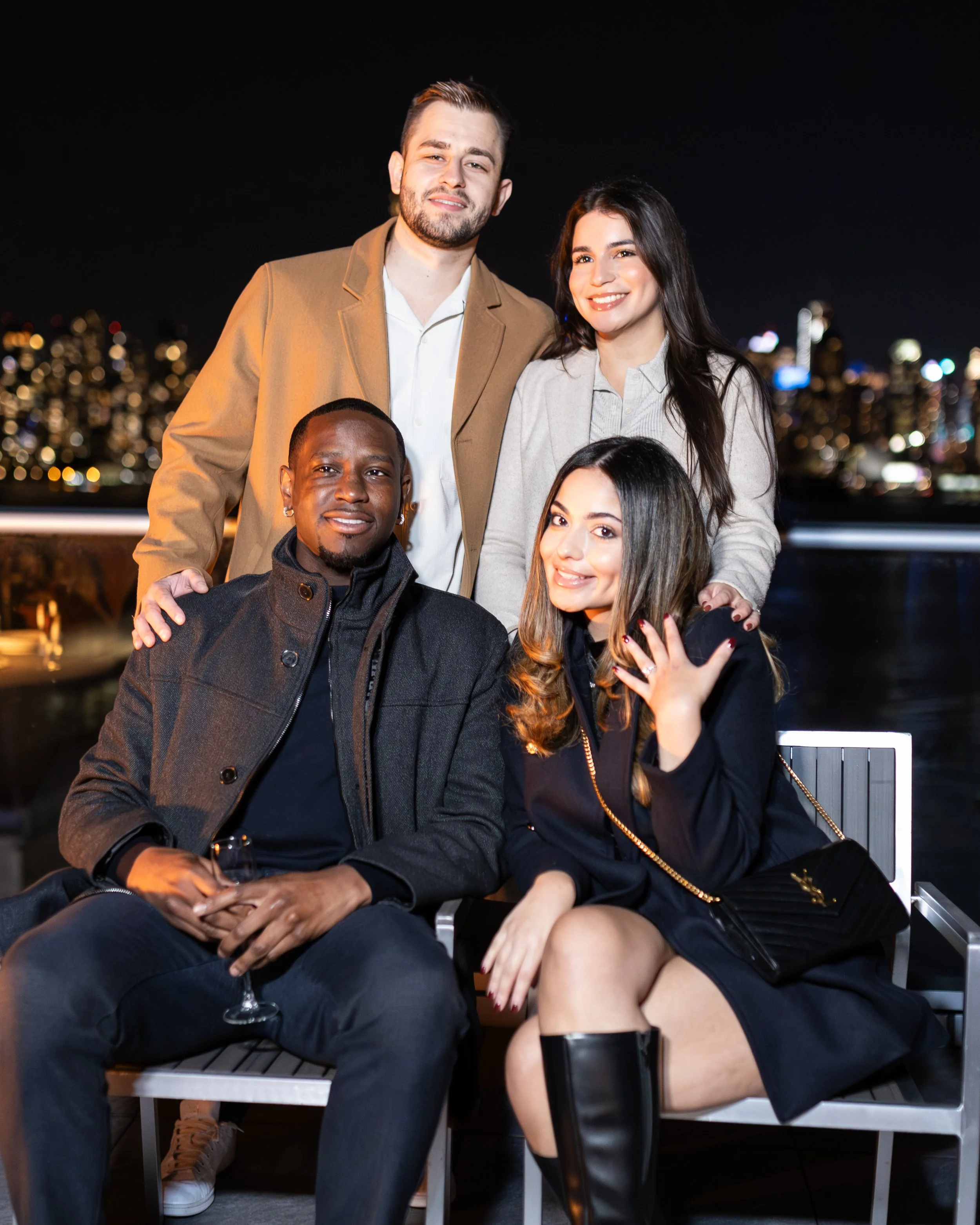 Group of five friends enjoying night out on a rooftop with city skyline background, some sitting and some standing, dressed casually.