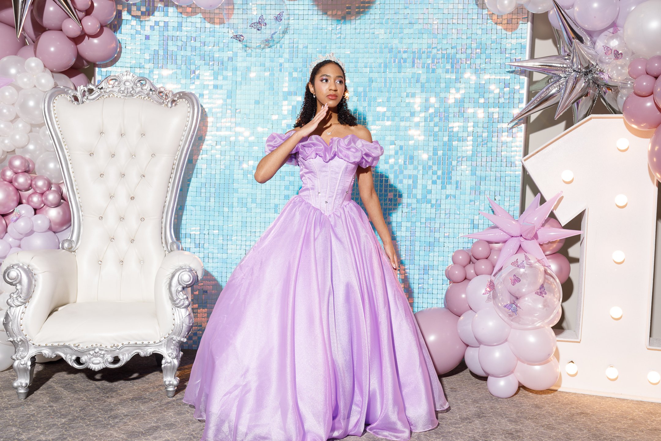 Young woman in a lavender ball gown standing in front of a blue mosaic backdrop with balloon decorations and a decorative chair.