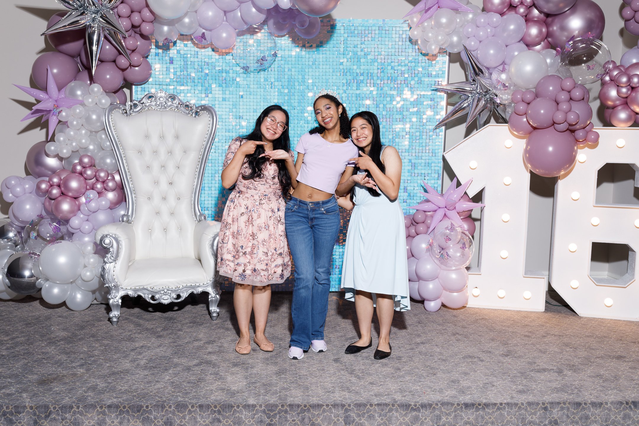 Three women smiling and posing together at a celebration or event, standing in front of a decorative backdrop with balloons, stars, and a mosaic wall, and a large white ornamental letter 'H' with lights.