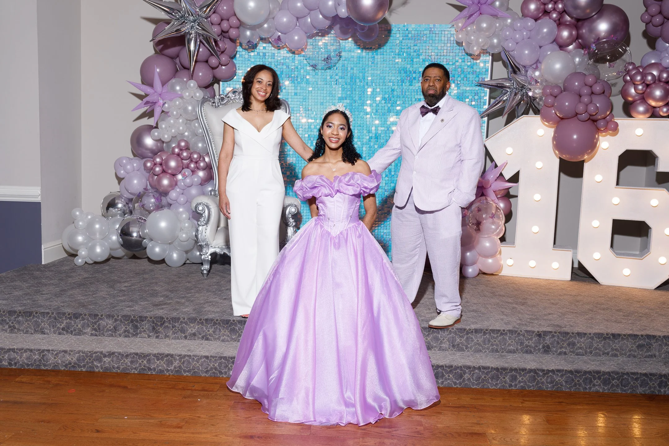 A girl in a purple ball gown is standing with two adults, a woman in a white jumpsuit and a man in a light-colored suit, in front of a balloon arch and a large number 16 sign, celebrating a special occasion.