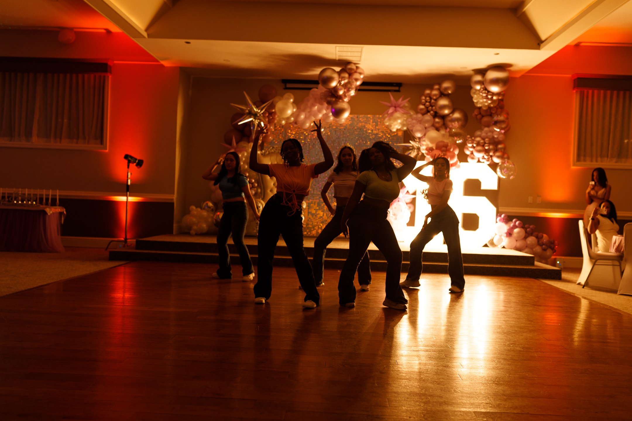 A group of people dancing on a wooden floor at a celebration or party, with colorful balloons and decorative elements in the background, and warm lighting.