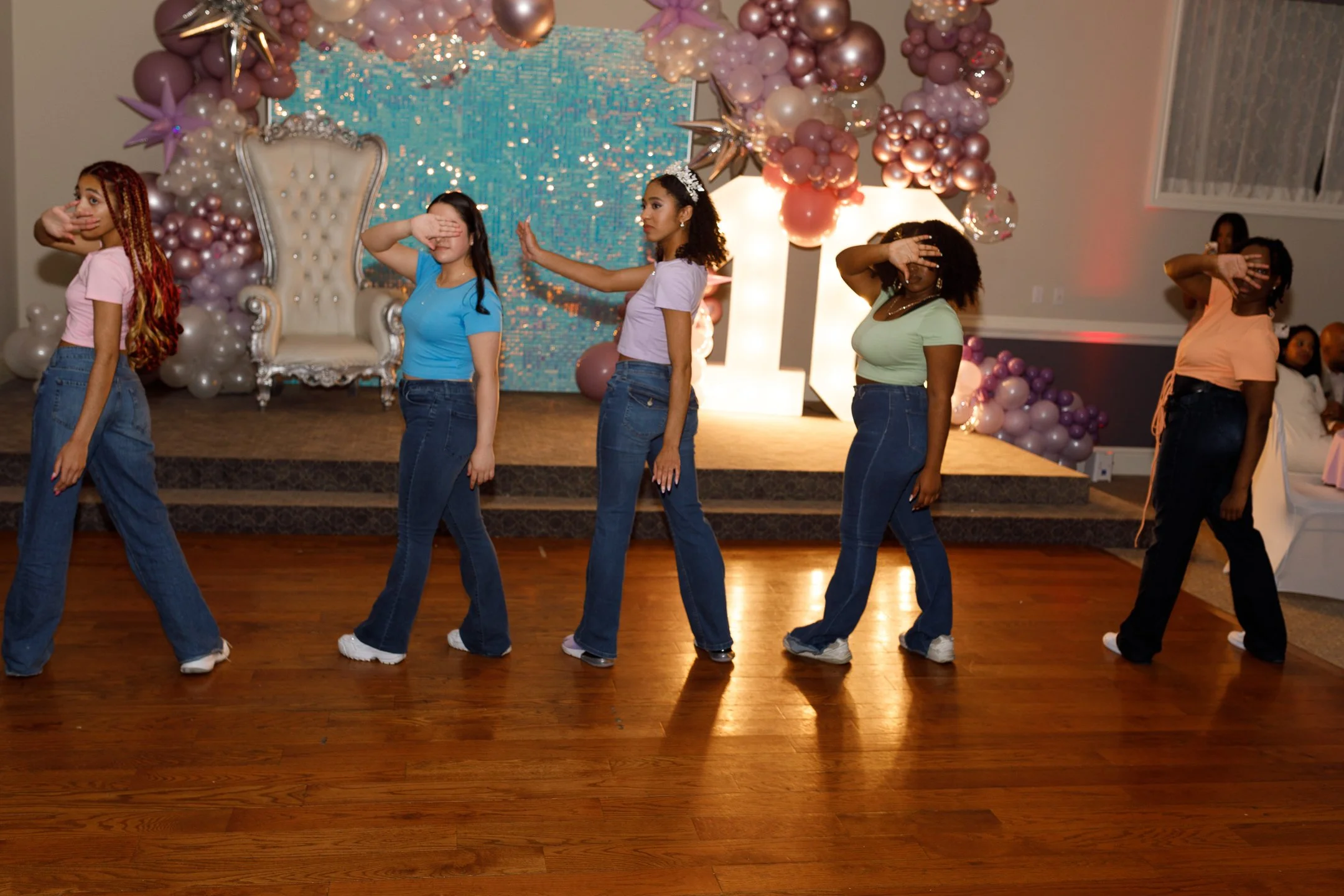 Six girls dancing in a line at an indoor event with a decorated stage in the background.