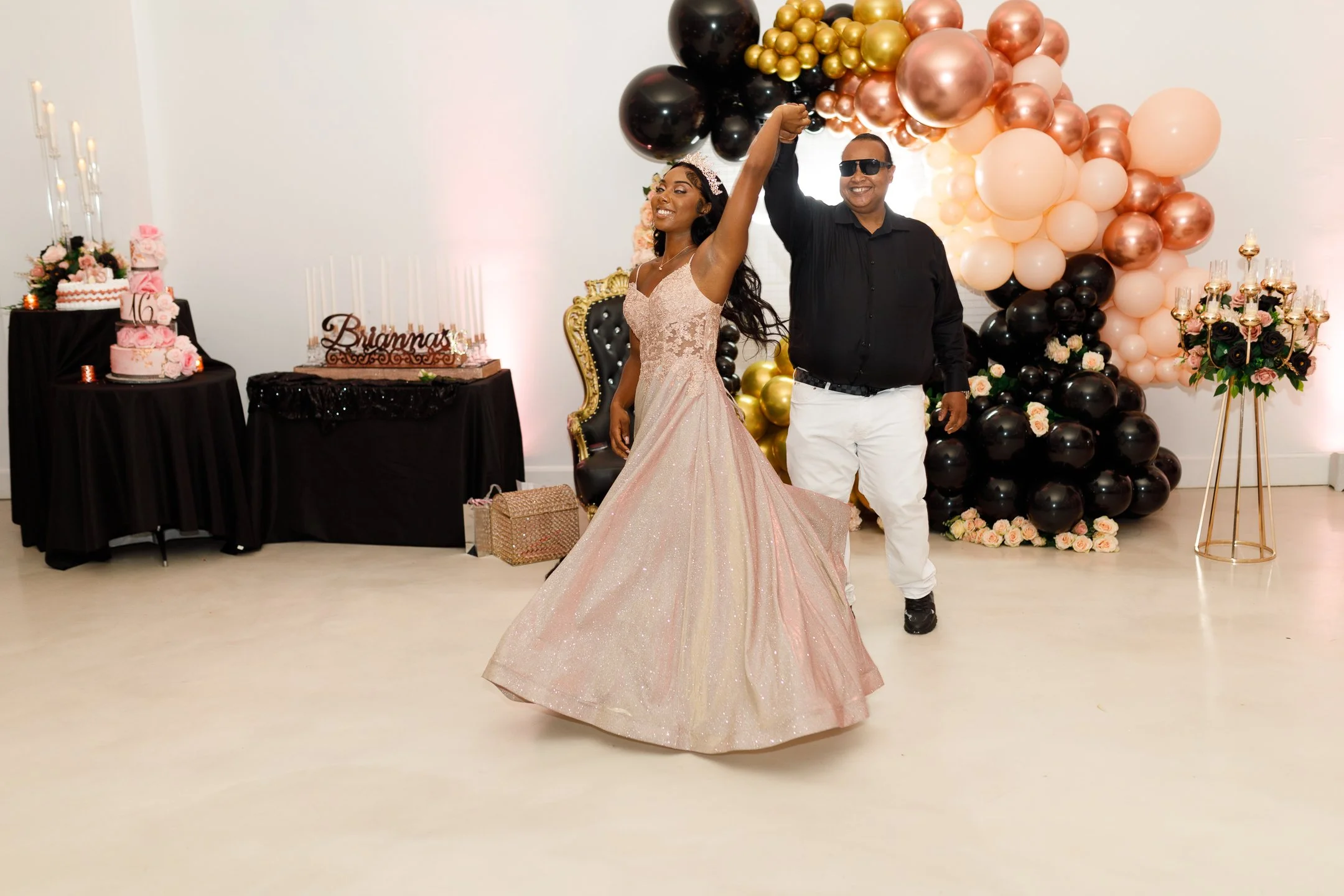 A young woman in a sparkling pink gown and a tiara dancing with a man in black shirt and white pants at a celebration with pink and black balloons and floral arrangements.