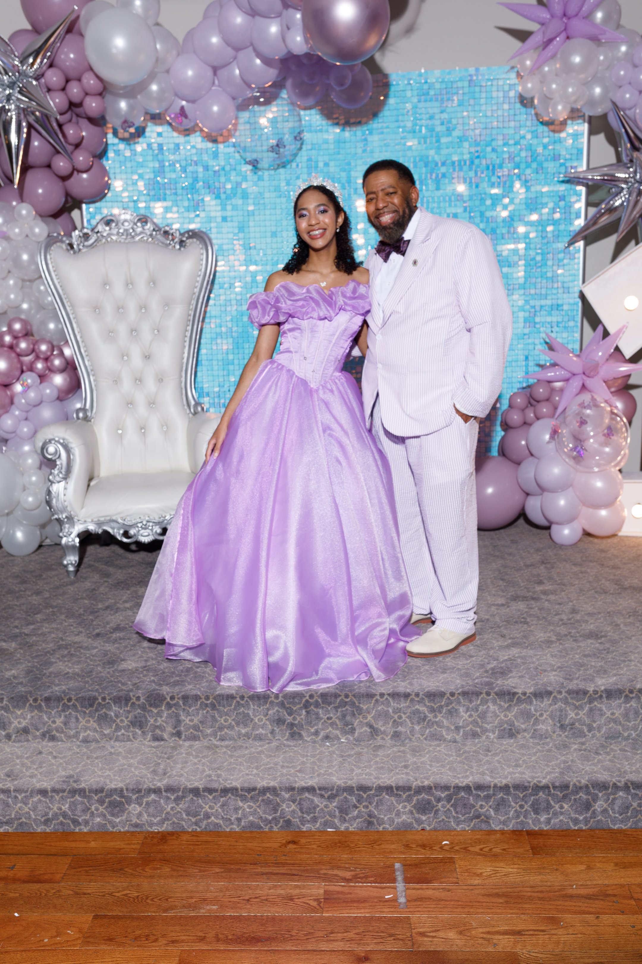 A young woman in a purple gown and an older man in a white suit with purple stripes standing together on a decorated stage with balloons and a blue mosaic backdrop, smiling.