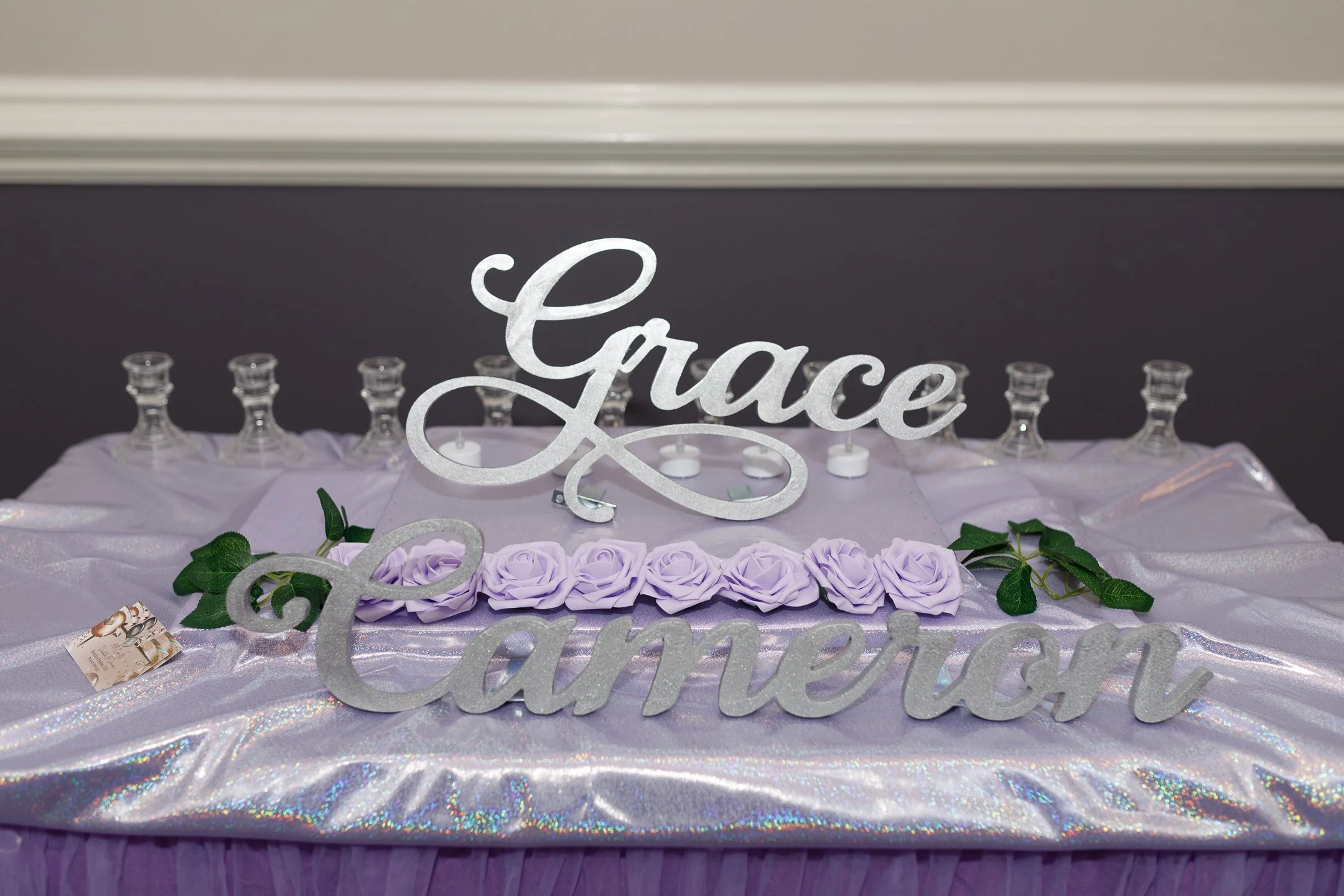 Decorative display with a large silver cutout of the name "Grace Cameron" on a table covered with shiny, lilac-colored fabric, purple artificial roses, green leaves, and glass candle holders.