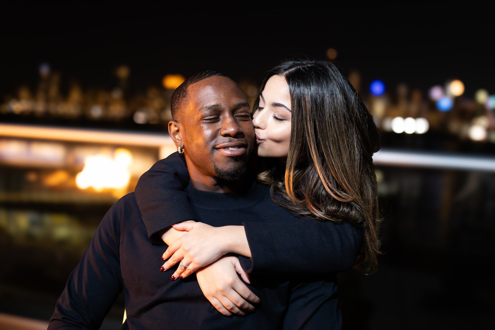 A woman with long dark hair kissing a man with short hair and a goatee on the cheek at night, with a cityscape and blurred lights in the background.