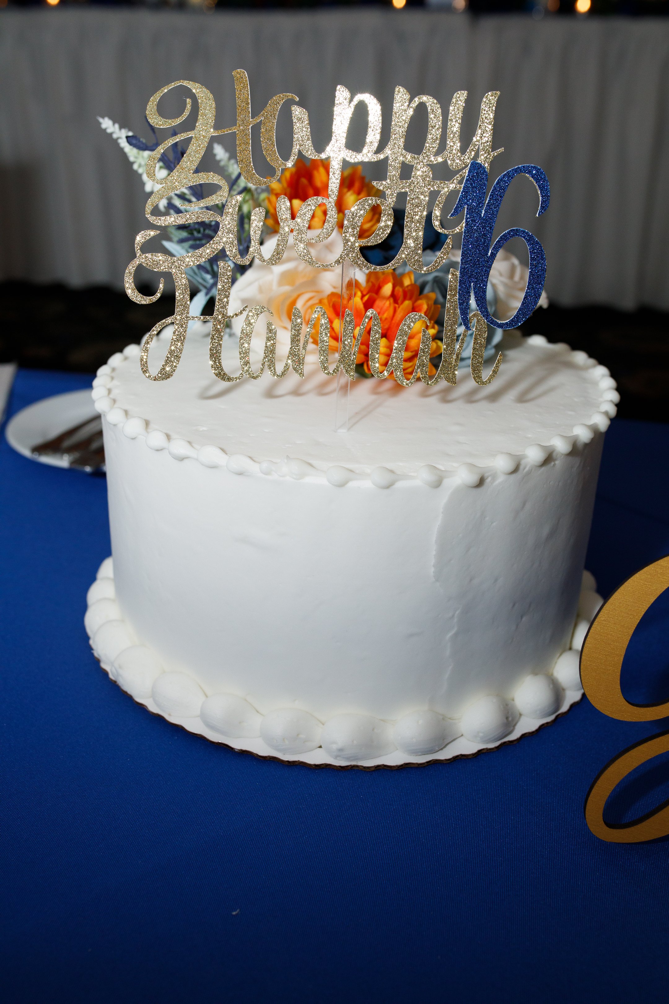 A round white cake with simple white icing decorated with a topper that says "happy sweet 16th Hannah" and orange flowers on top, placed on a blue tablecloth.