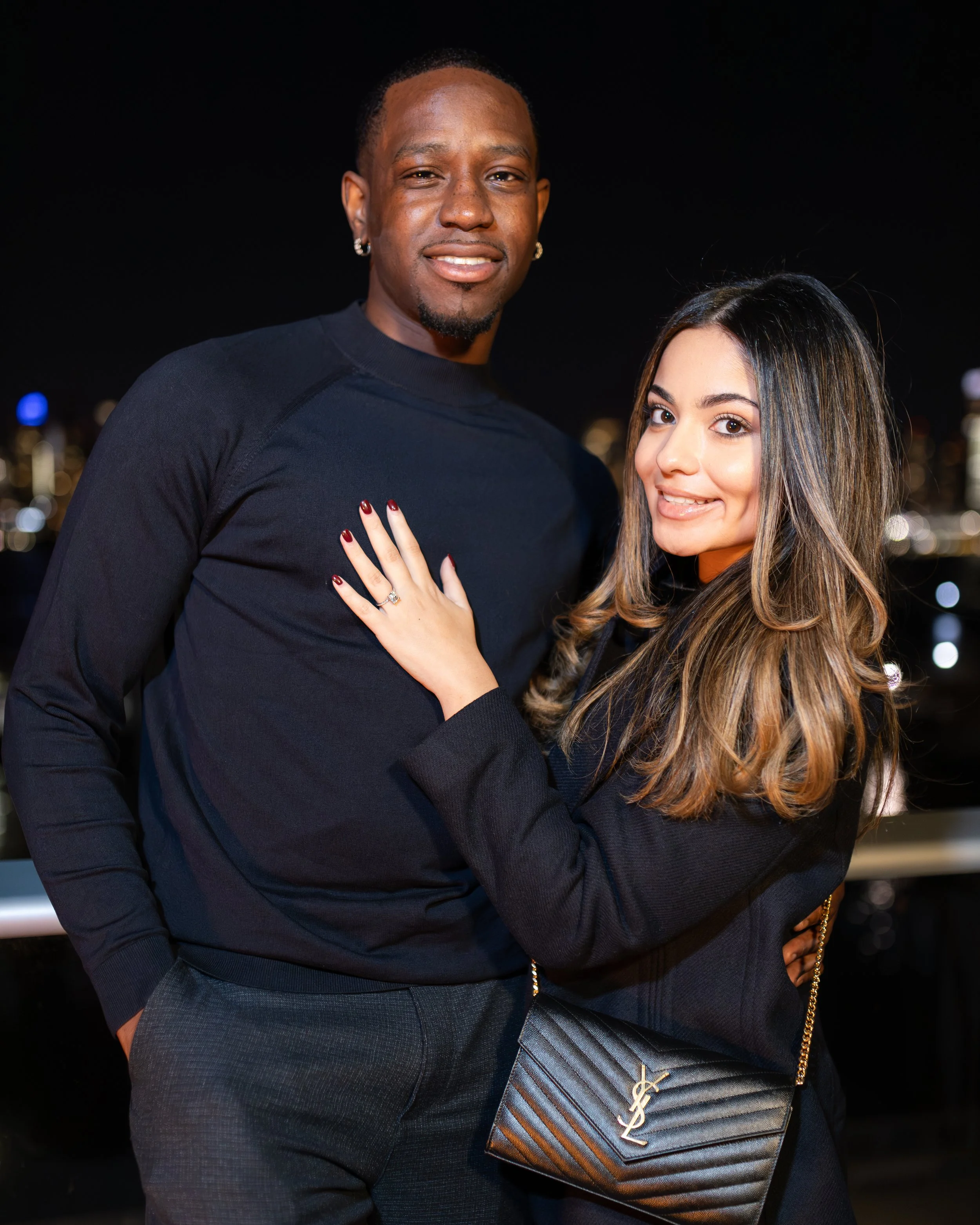 A man and woman posing together at night with city lights in the background, the woman has her hand on the man's chest and is holding a YSL purse.