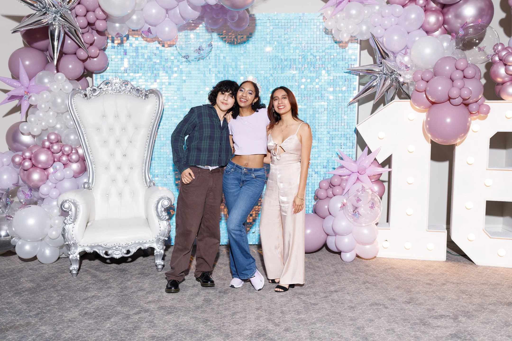 Three women standing together at a decorated event, with pink and white balloons, star-shaped balloons, a large illuminated letter 'E', and a sparkling blue backdrop.