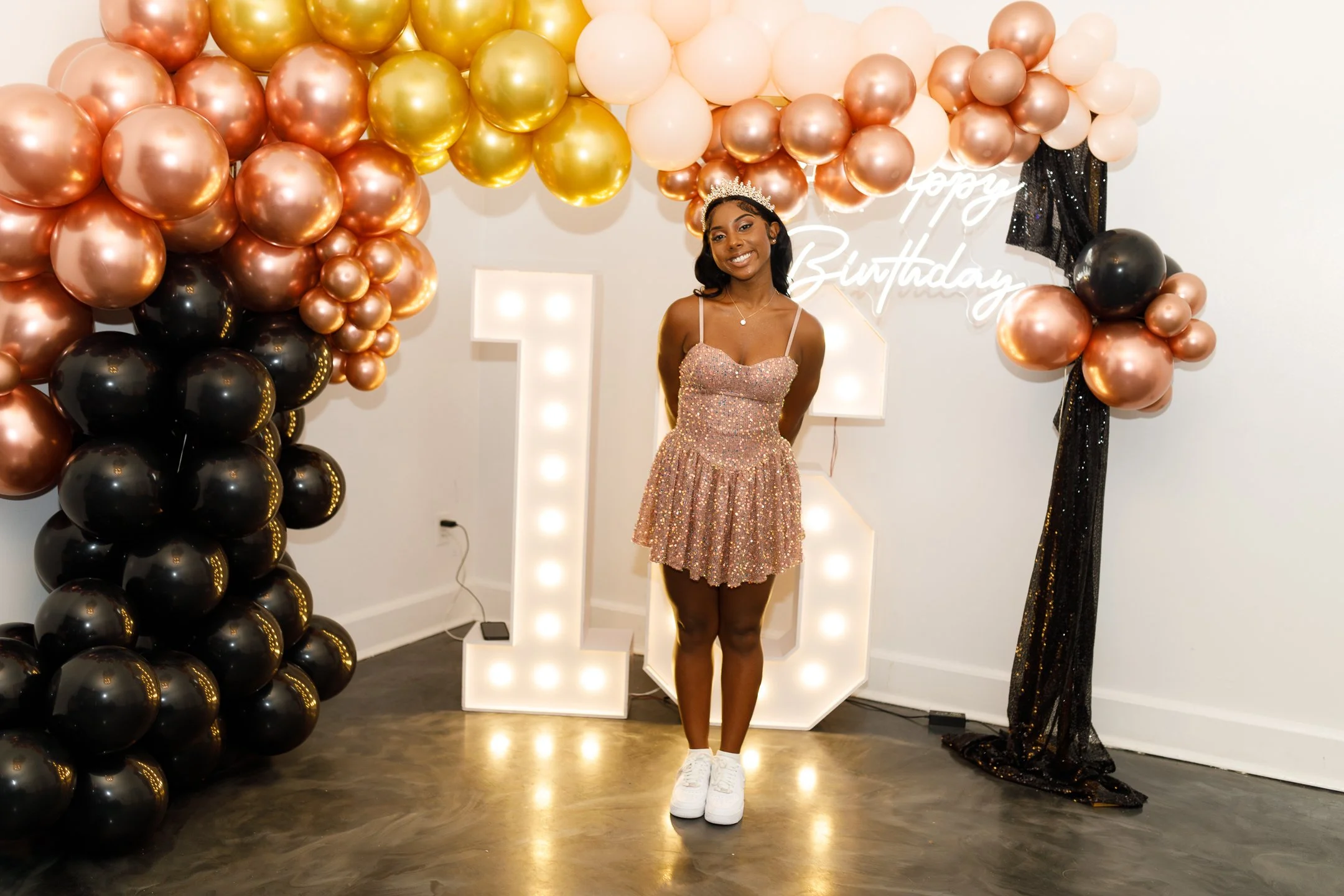 Young woman standing in front of a birthday decoration with pink, black, gold, and pearl balloons, a lit '1' sign, and a neon 'happy birthday' sign. She is wearing a sparkly dress and a tiara.