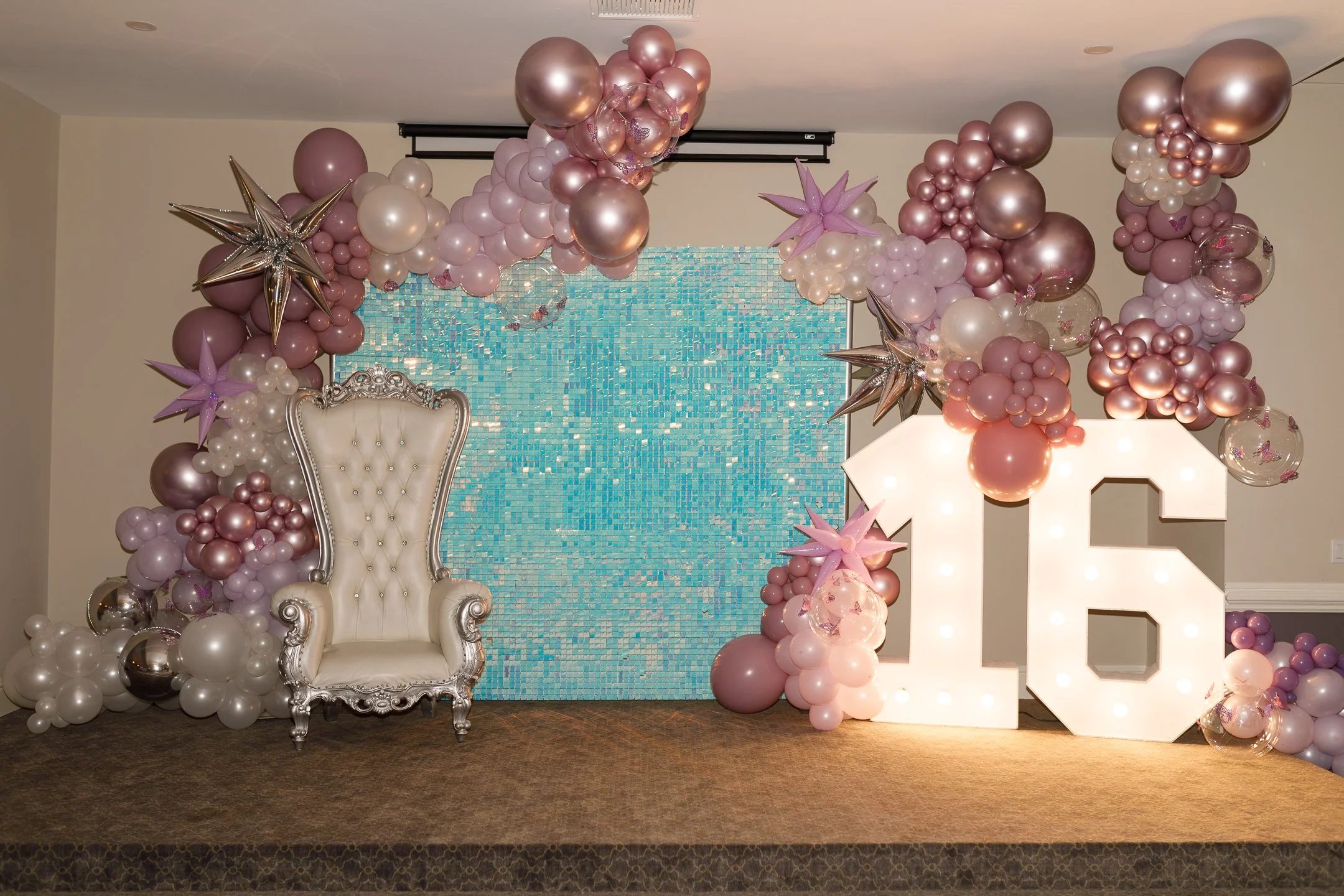 Decorative photo with a white ornate chair, a large illuminated number 16, and pink, white, and metallic balloons arranged in an arch around a blue tiled backdrop, likely for a 16th birthday celebration.