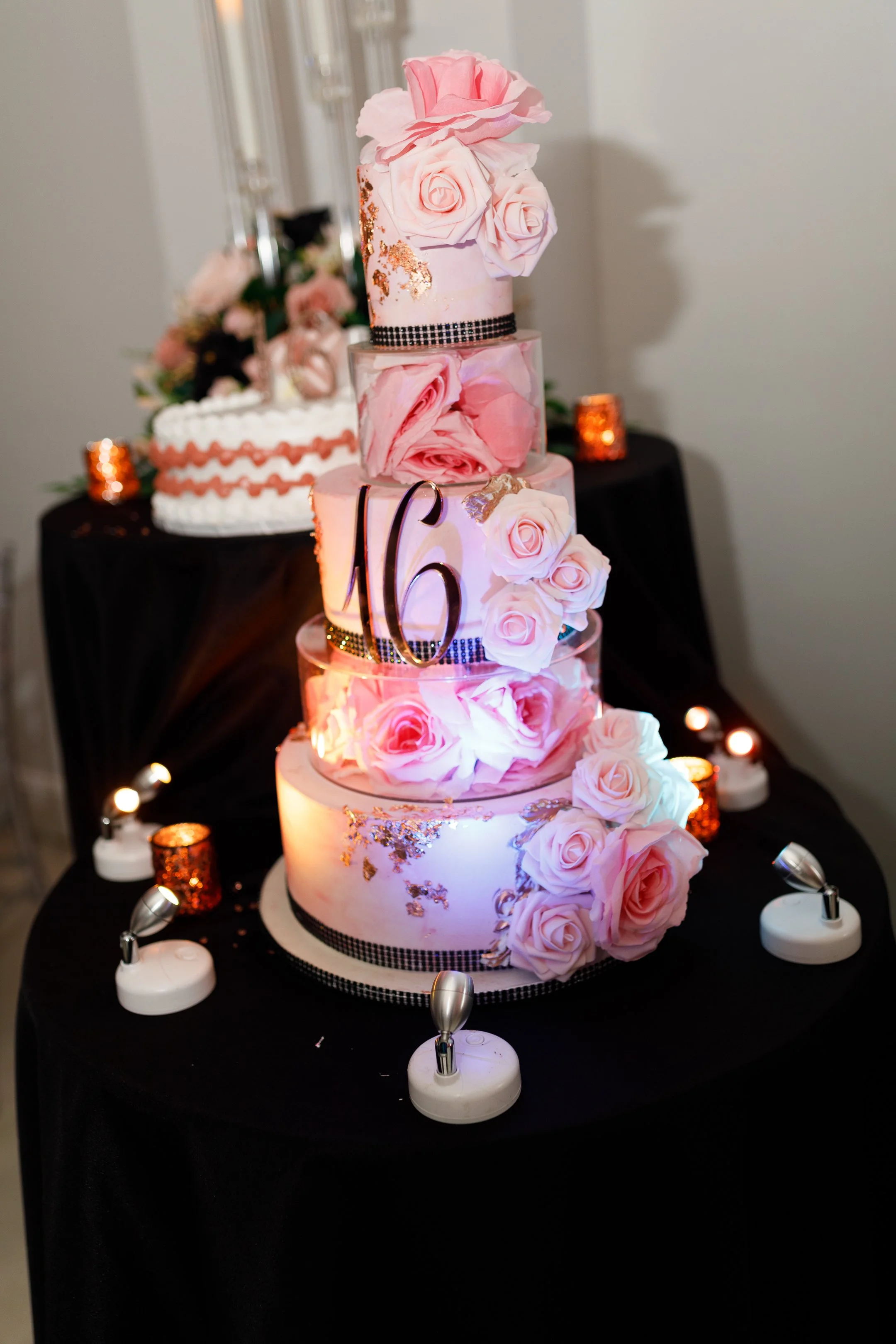 A pink tiered wedding cake decorated with pink roses, gold leaf accents, and black ribbon details, numbered '16', surrounded by small decorative lights and candles on a black table.