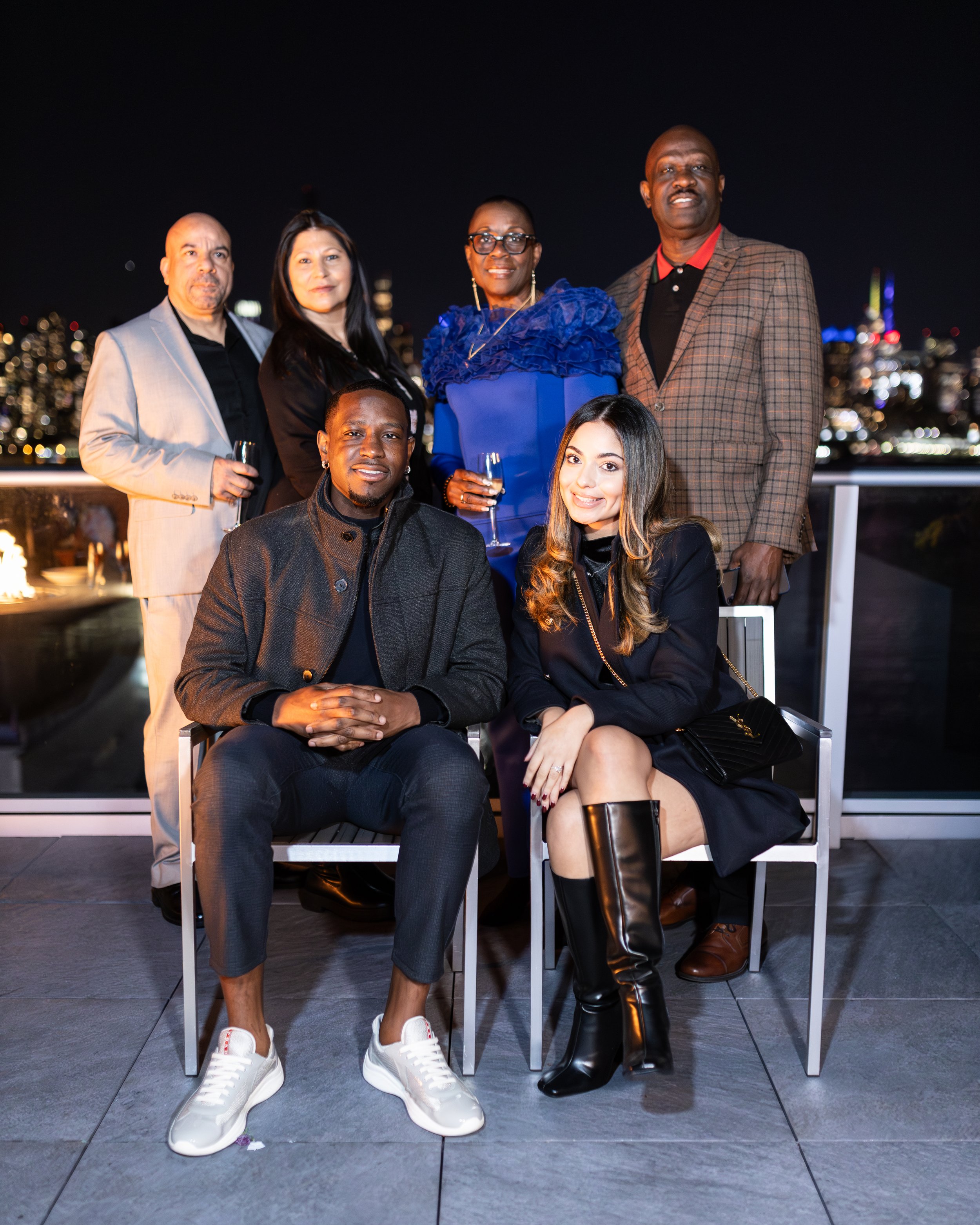 Group of six people at a nighttime rooftop gathering with city skyline in the background, some holding champagne glasses, dressed in formal and semi-formal attire.