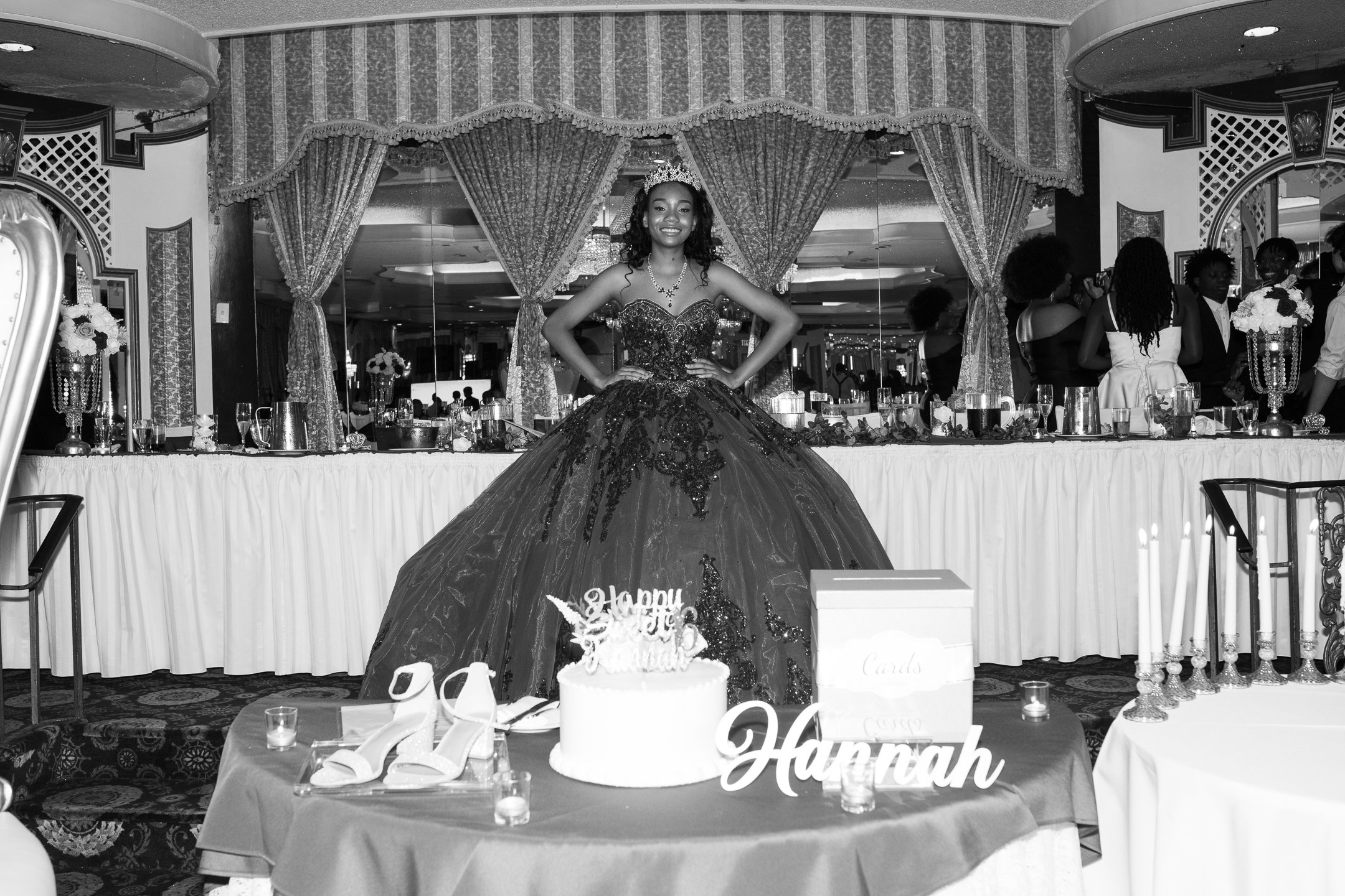A young woman in an elegant ball gown stands with her hands on her hips, smiling at a celebration event. She is wearing a tiara and jewelry, and behind her is a decorated banquet table with candles, flowers, and glasses. In the foreground, there is a
