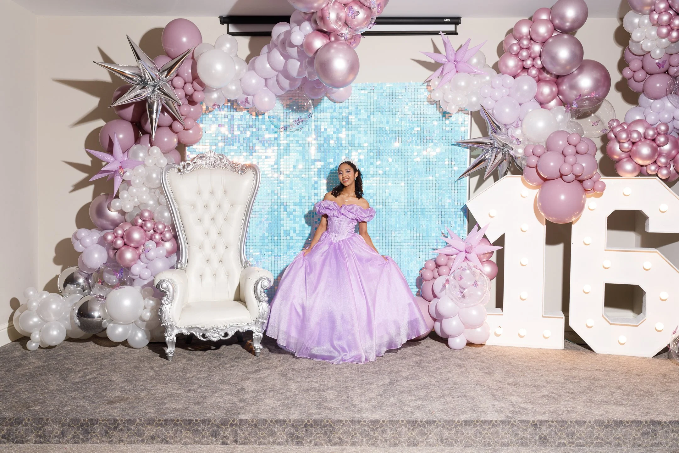Young woman in a purple ball gown standing on a decorated stage with pink, white, and silver balloons, a white ornate throne chair, and large illuminated letters 'LOVE' in the background.