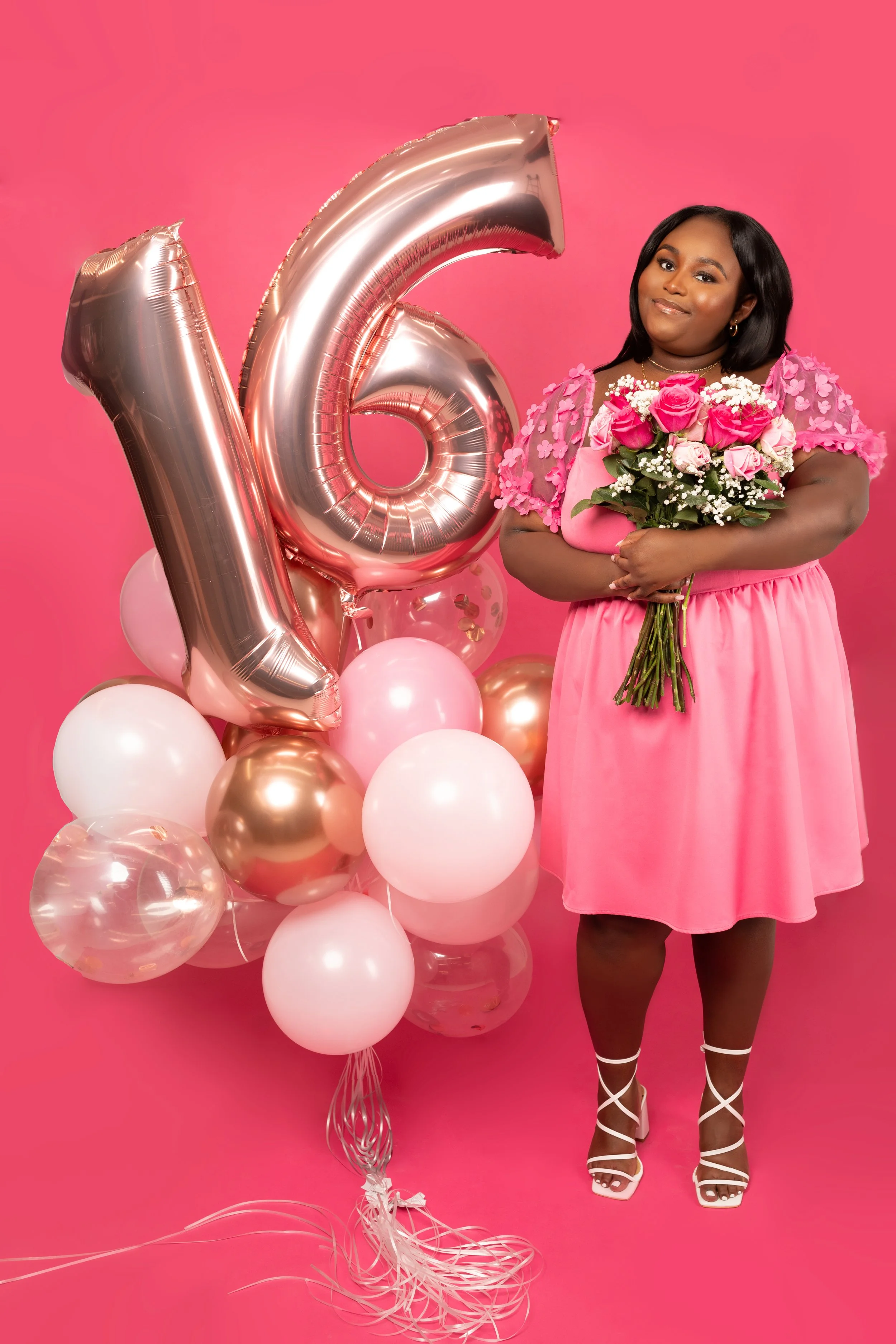 A woman in a pink dress holding a bouquet of pink and white roses, standing next to large pink, white, and gold balloons, with a pink background.