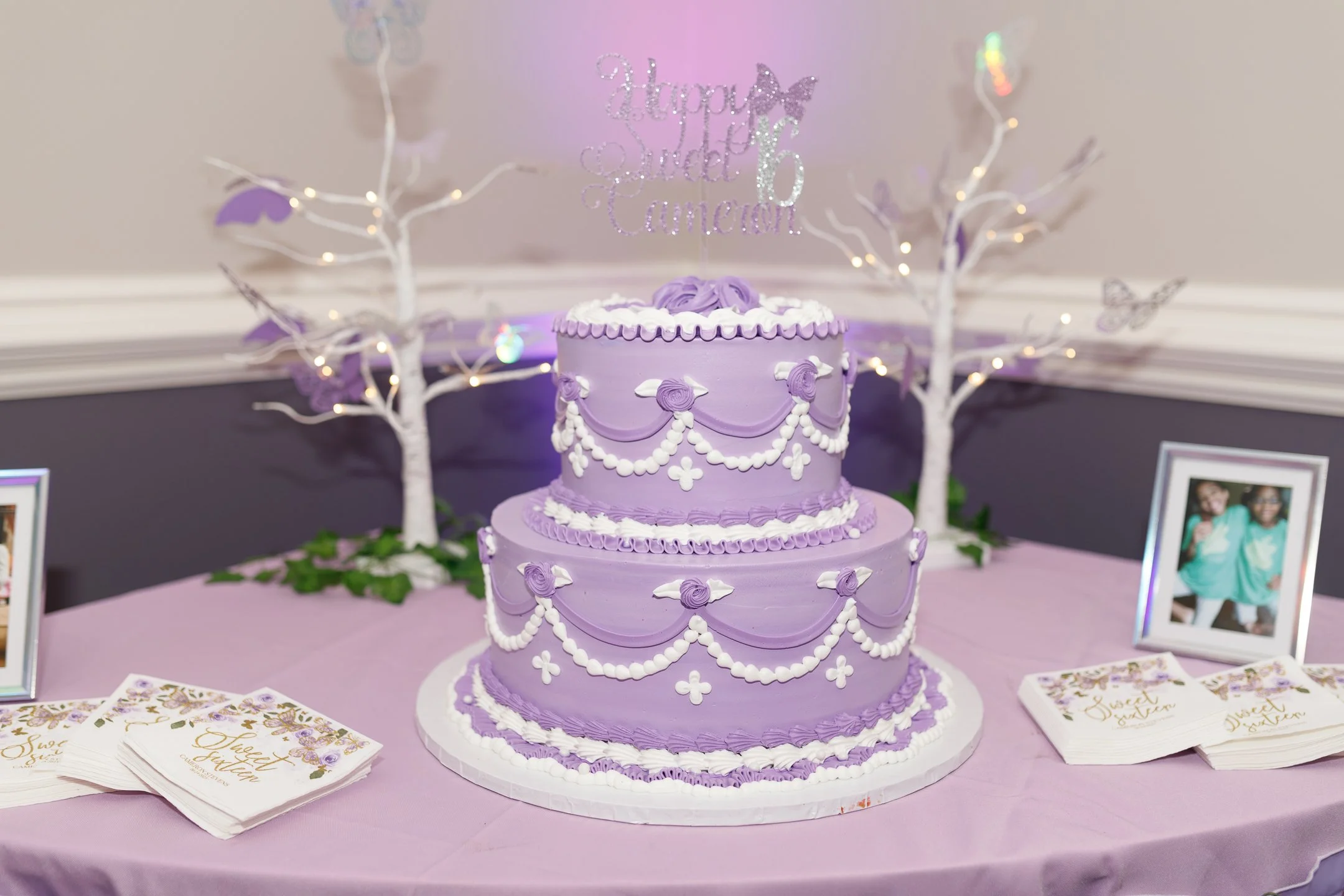 Two-tier lavender birthday cake decorated with white and purple icing swags, roses, and pearls, topped with a glittery 'Happy Sweet Sixteen' topper. The cake table has framed photos, pink and purple brochures, and is decorated with white paper trees 