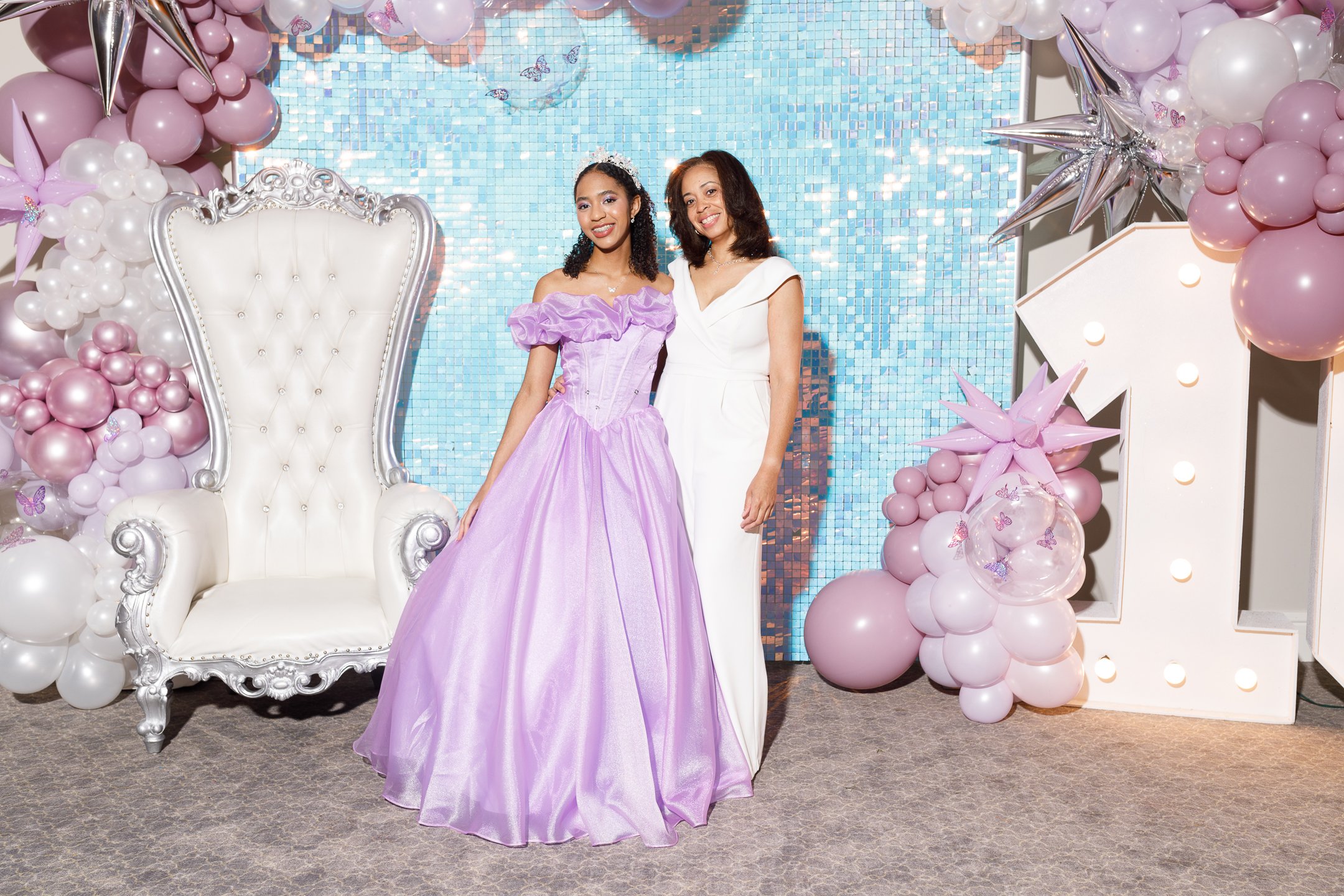 Two women standing at a celebration with a pink and white balloon decoration, a large white chair, and a mosaic blue background. One woman is wearing a purple dress, and the other a white dress.