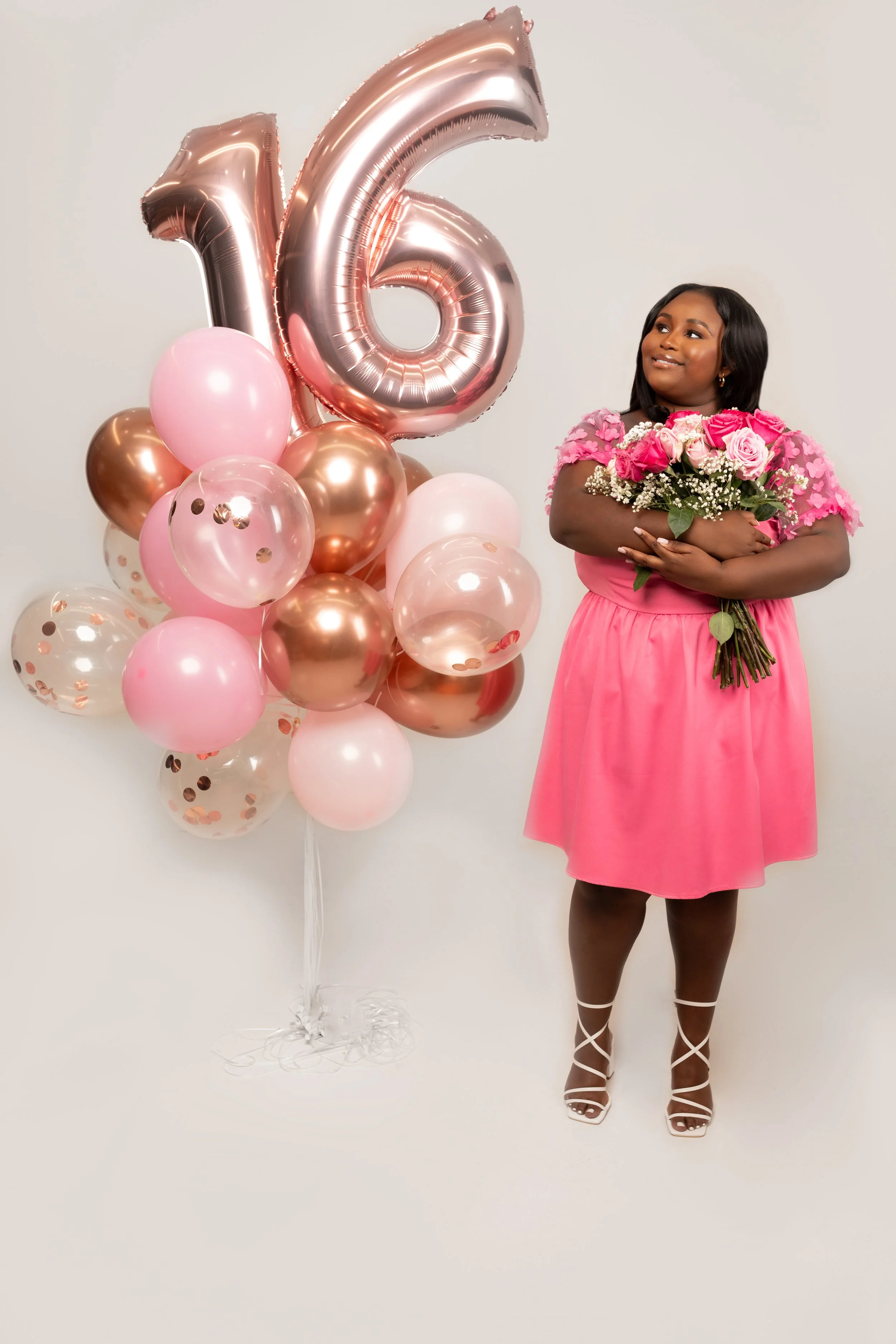 Woman holding a bouquet of pink roses standing next to a large '16' balloon with pink, white, and metallic balloons in the background.