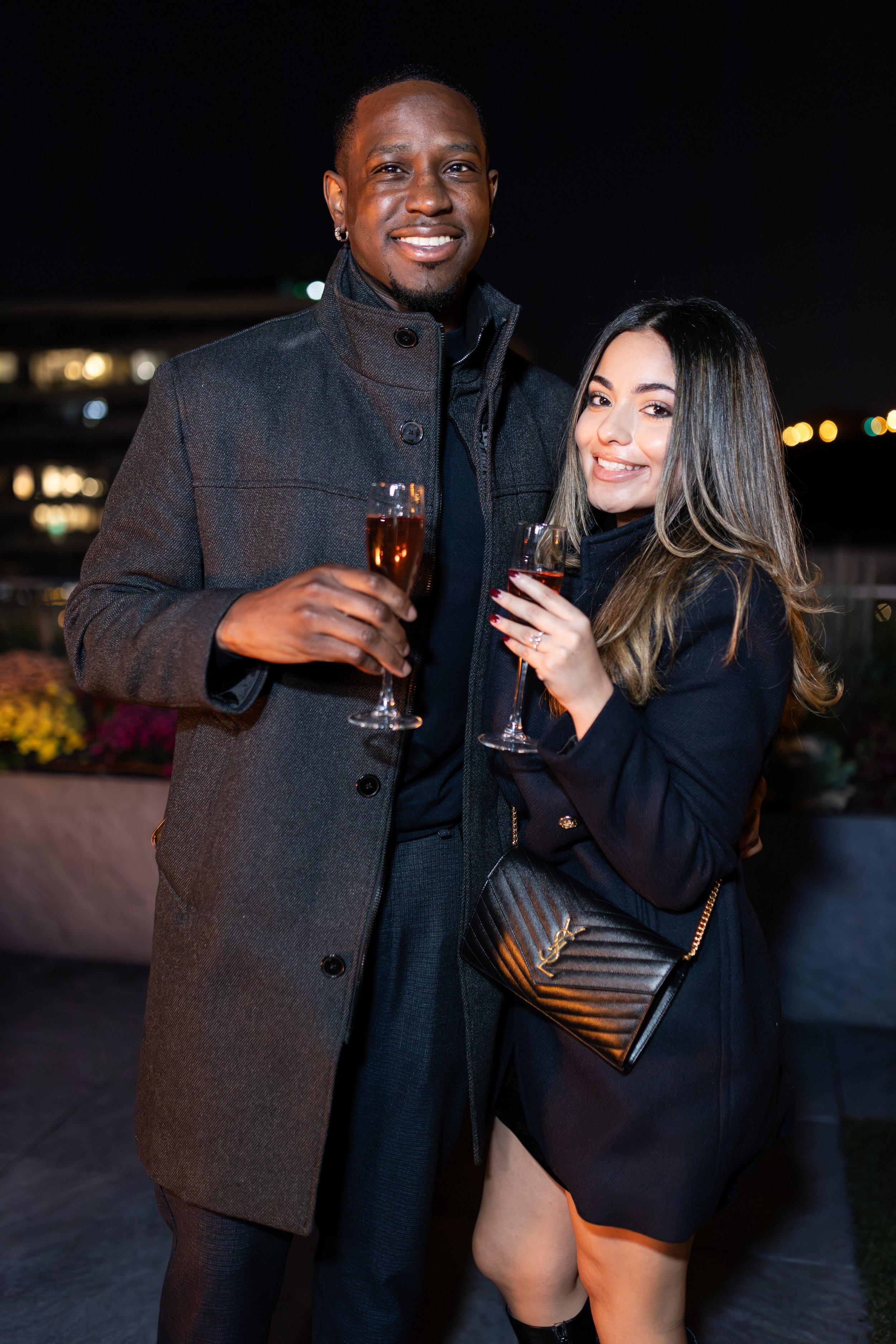 A man and woman dressed in evening attire holding glasses of champagne at a nighttime outdoor event.