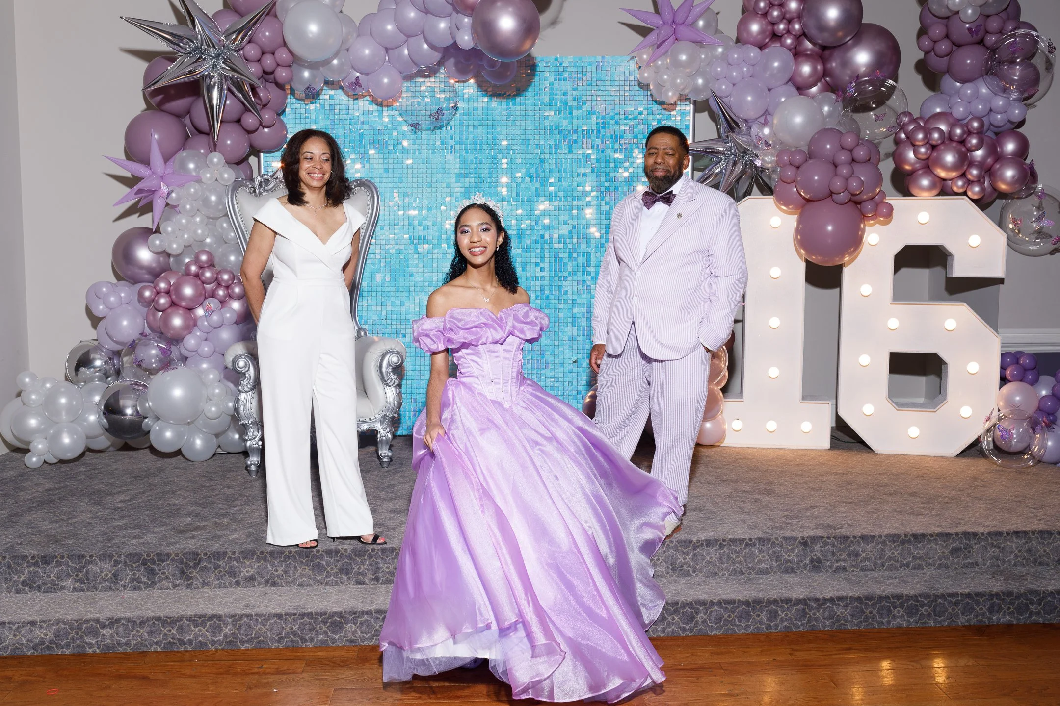 A young woman in a purple ball gown sitting on a chair, flanked by two adults in formal attire, at a celebration with balloon decorations and large lit-up numbers '16' in the background.