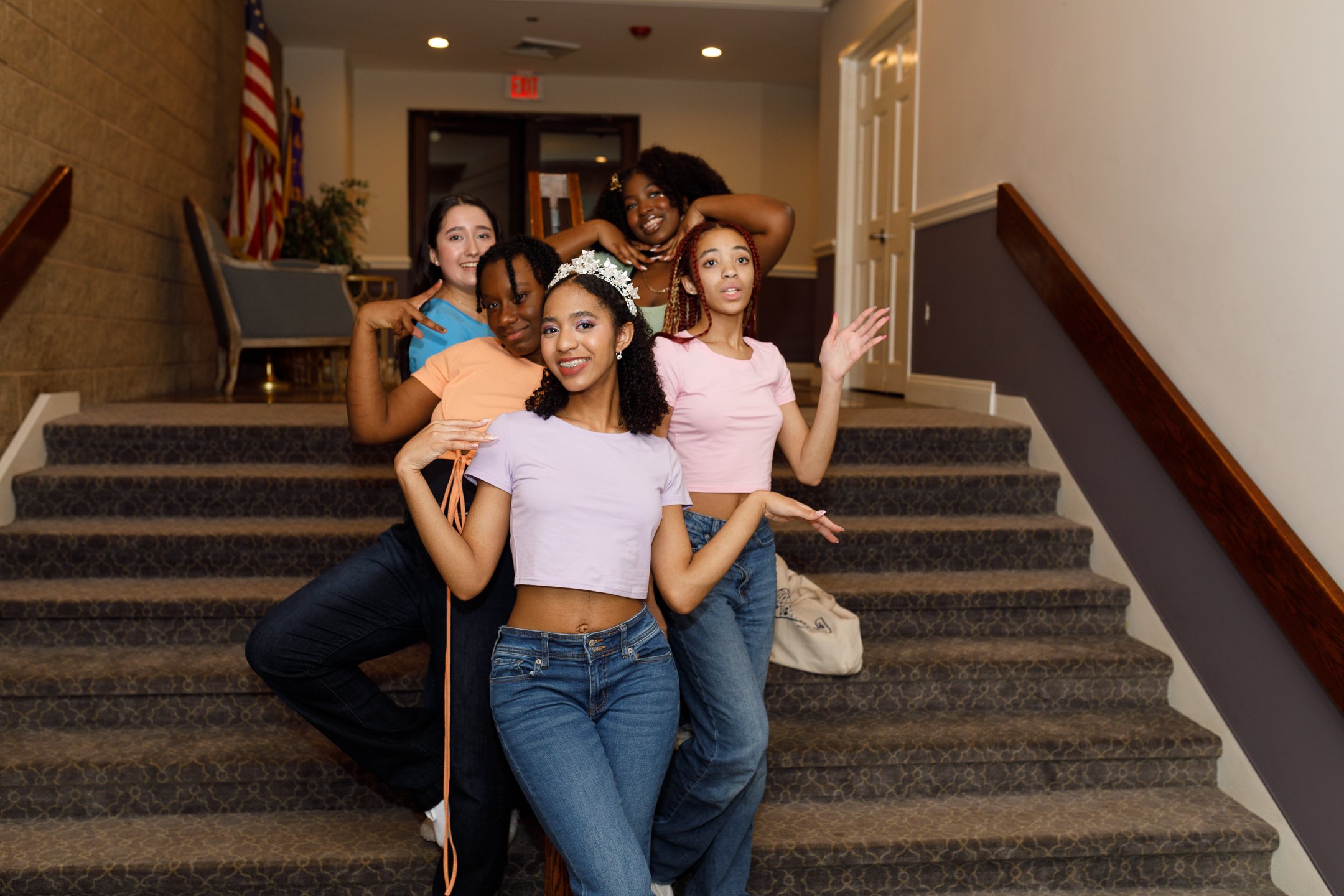 Group of six young girls posing on a staircase indoors, smiling and making gestures.