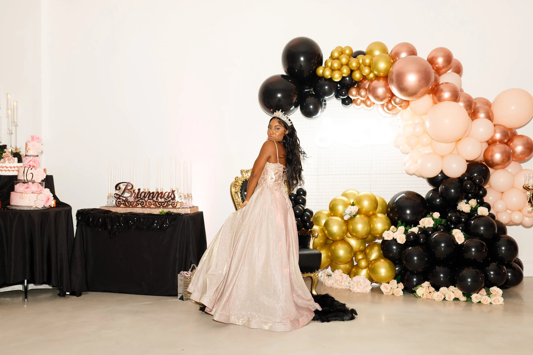 A young woman in a light pink, sparkly gown and a crown, standing in front of a balloon arch with black, gold, and pink balloons, at a celebration event.