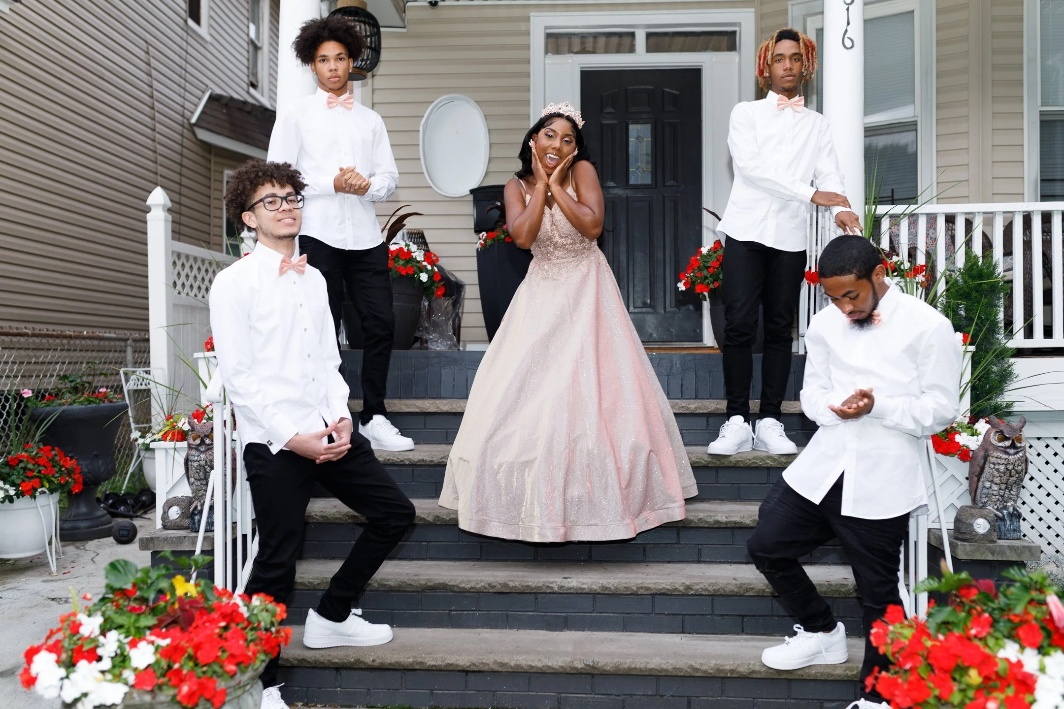 A young woman in a pink ball gown and tiara standing on stairs in front of a house, smiling and posing happily with her hand on her cheek. Five young men dressed in white shirts, black pants, and pink bow ties surround her, three standing on the stai