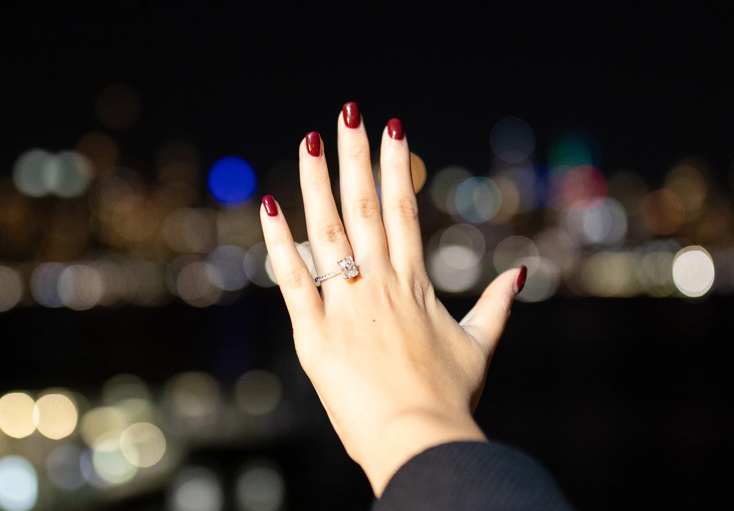 A woman's hand with a diamond engagement ring shows off her nails painted red against a blurred cityscape at night.