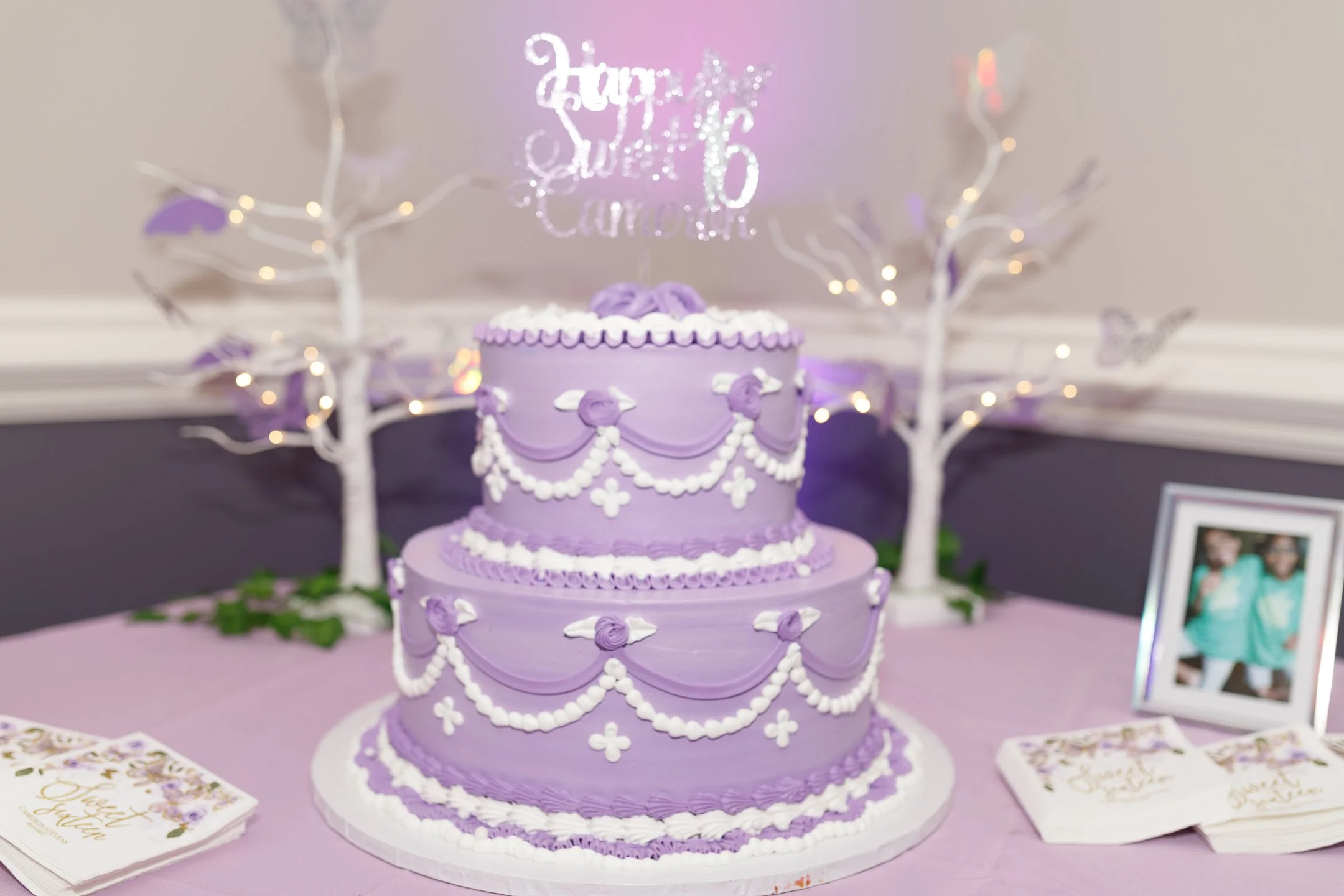 A two-tiered purple birthday cake decorated with white icing swags, dots, and floral accents, with a glittery sign on top reading 'Happy 16th Sweet 16 Birthday.' The cake is on a table with pink tablecloth, flanked by white decorative trees with ligh