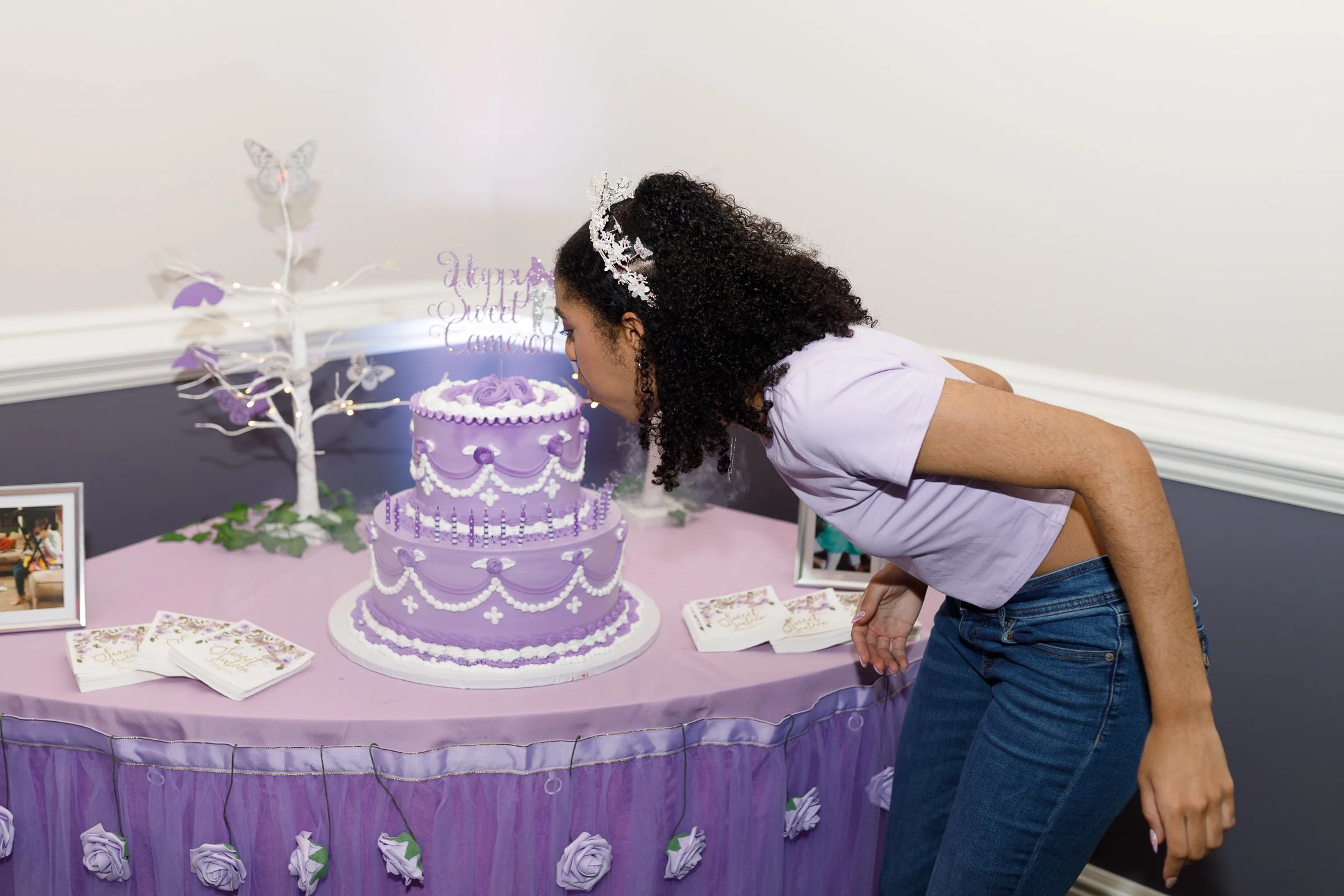 A woman leaning forward to blow out candles on a purple birthday cake decorated with white icing, flowers, and pearls, with a purple and white themed birthday party table in the background.