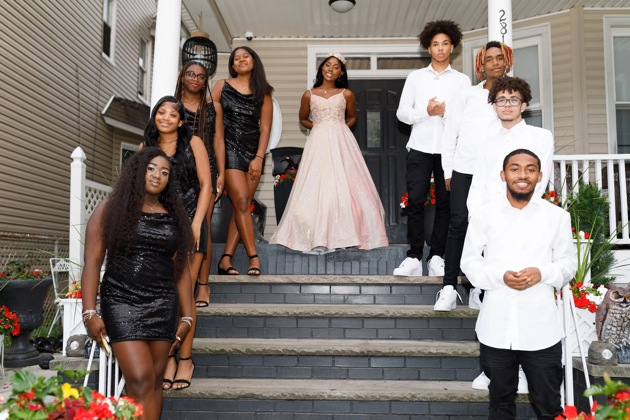 A group of ten young people, dressed in formal attire, standing on the front steps of a house decorated with flowers, with some wearing black dresses, some in white shirts and black pants, and one girl in a pink gown, smiling and posing for a photo.