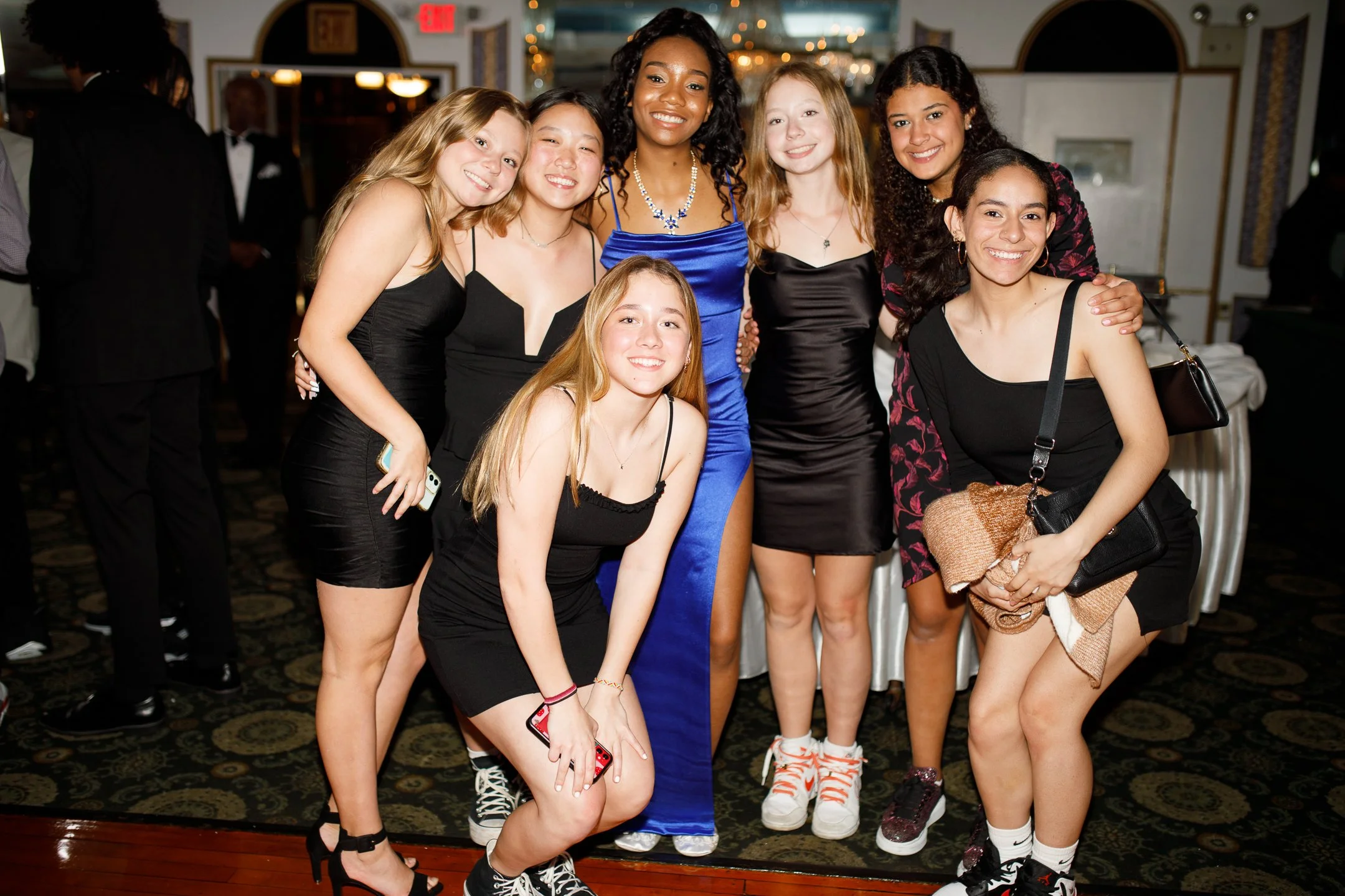 Group of seven young women at a formal event, smiling and posing together indoors with a dark patterned carpet and illuminated background.